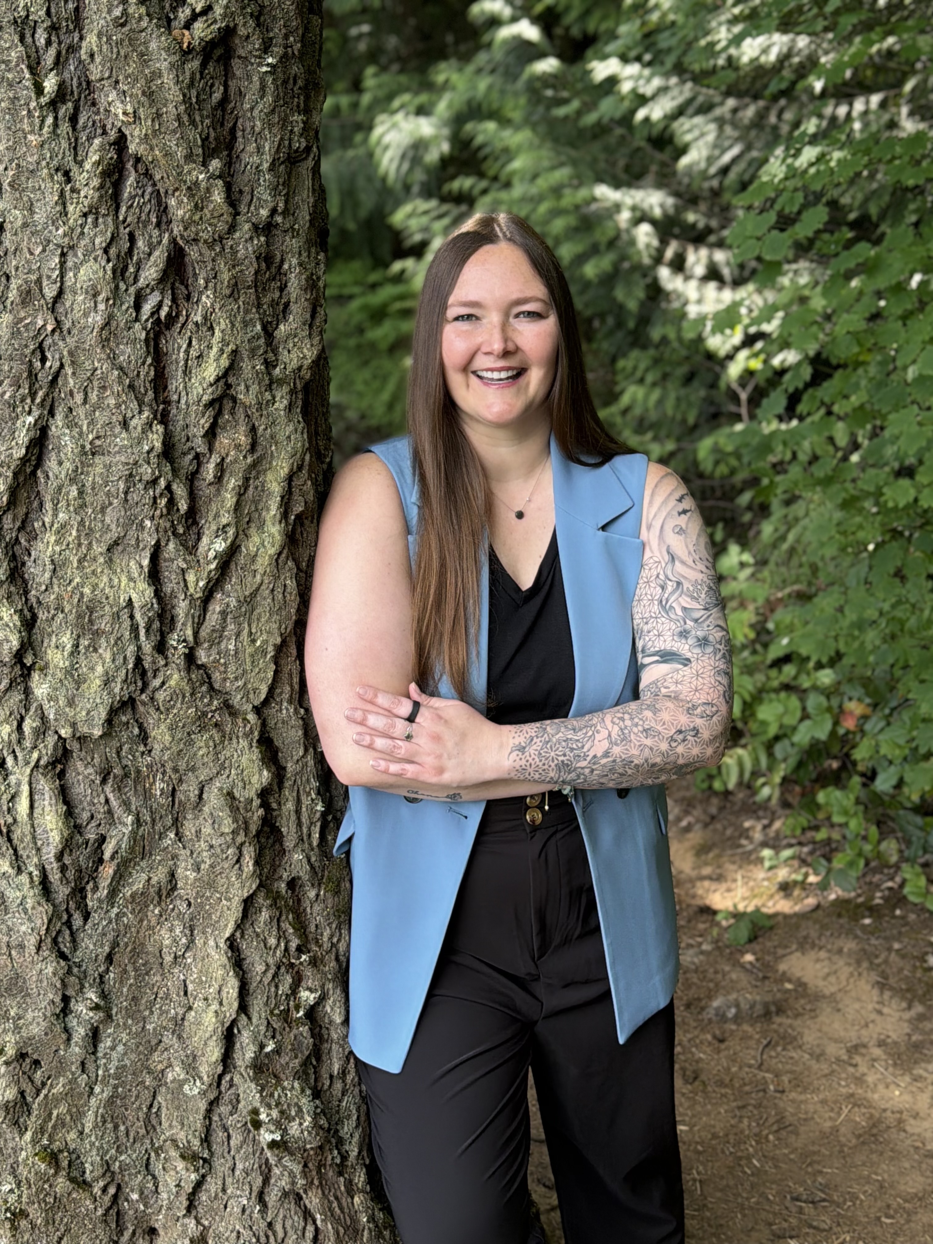 Ashley Hass, smiling woman with long hair and tattoos on arm leaning against a large tree outdoors.