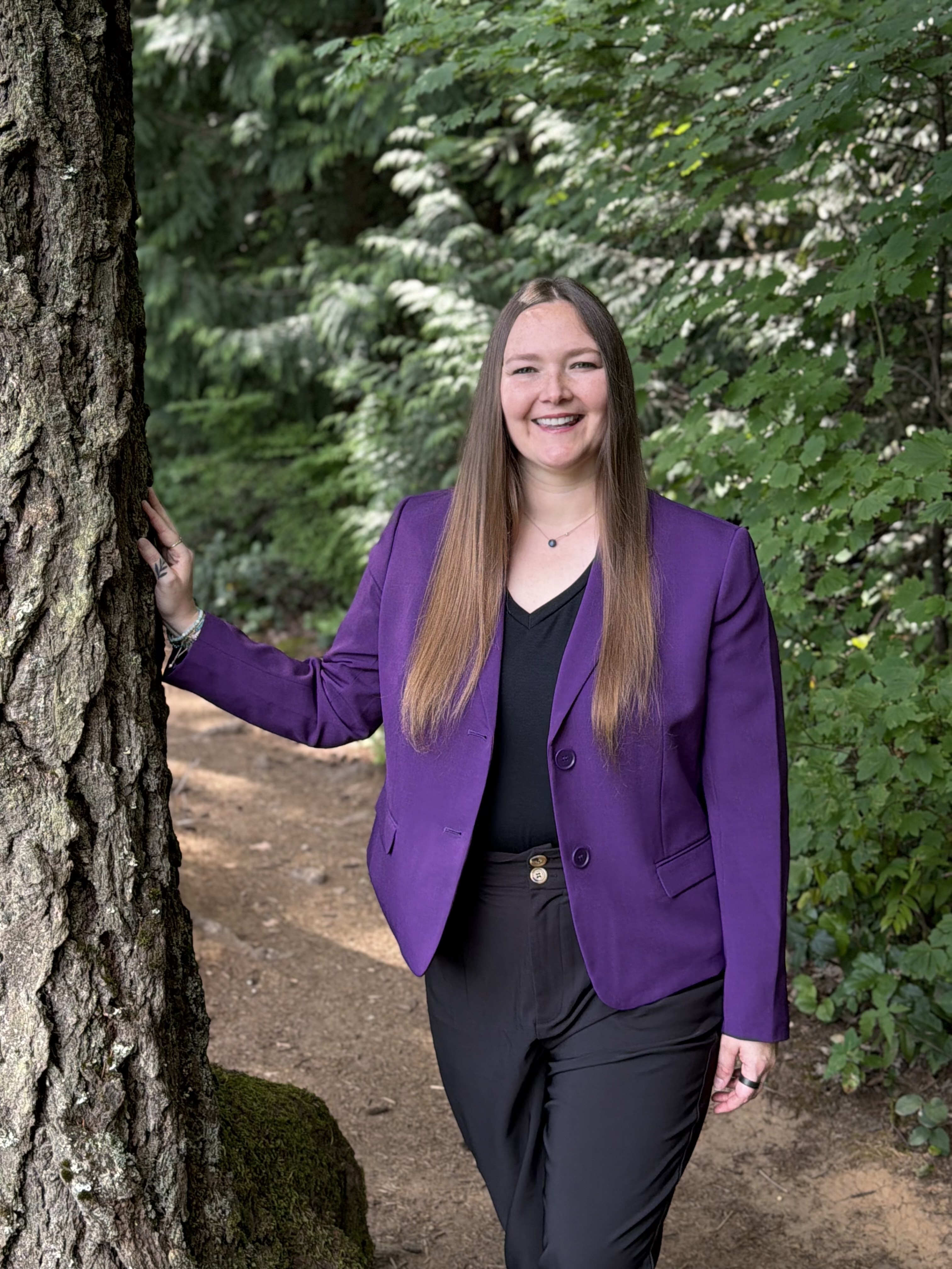 Smiling woman in purple blazer and black pants standing by a tree in a forest setting.