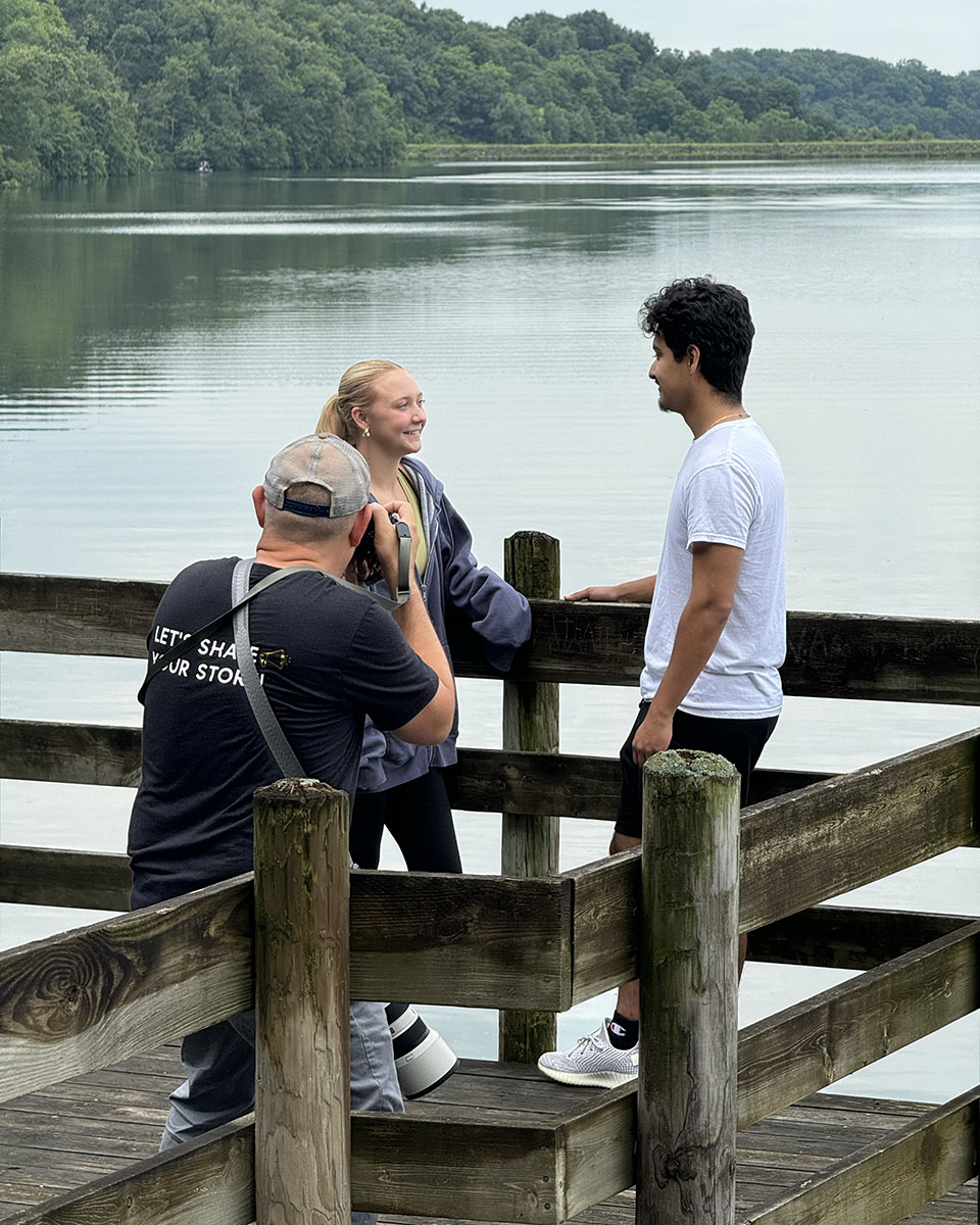 A Skorynko Media Group photographer captures two people posing on a wooden dock overlooking a lake, surrounded by lush trees.