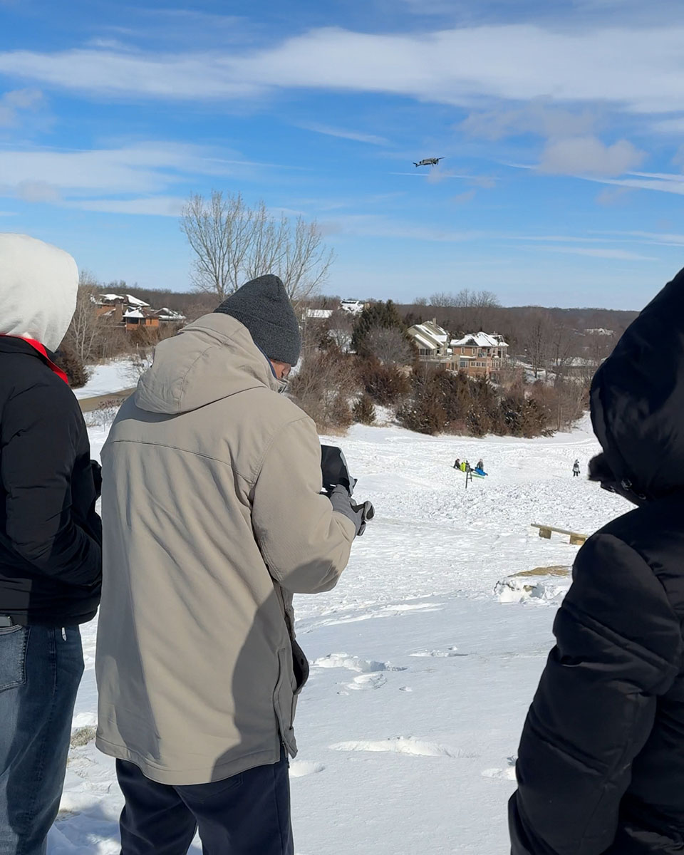 Group of people in winter jackets filming a drone flying over a snowy hill where others sled. Snow-covered houses and trees in the background under blue sky.