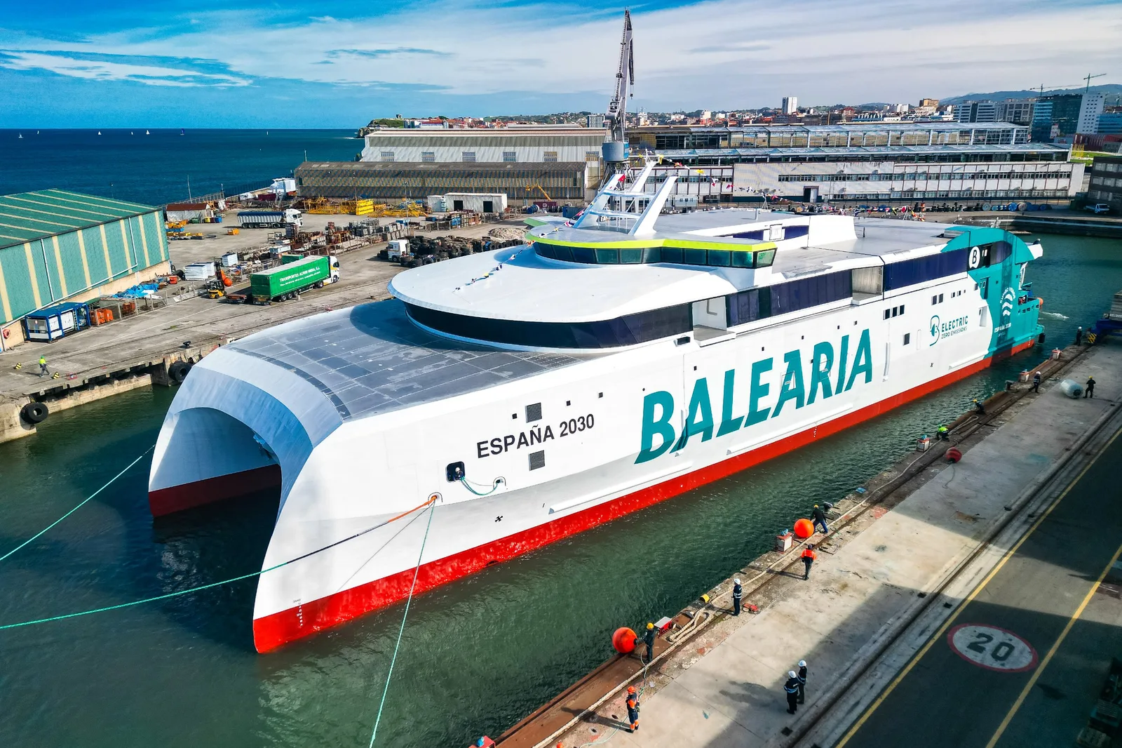 Vue aérienne du catamaran électrique de Baleària, coque blanche et rouge marquée España 2030, amarré dans le bassin du chantier Armón de Gijón après sa mise à flot.