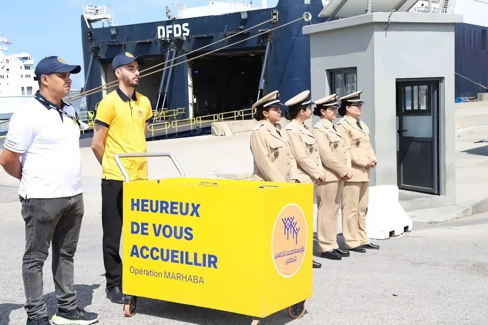 Two Marhaba staff members and four officers in beige uniforms stand by a yellow cart reading “Heureux de vous accueillir,” in front of a DFDS ferry.