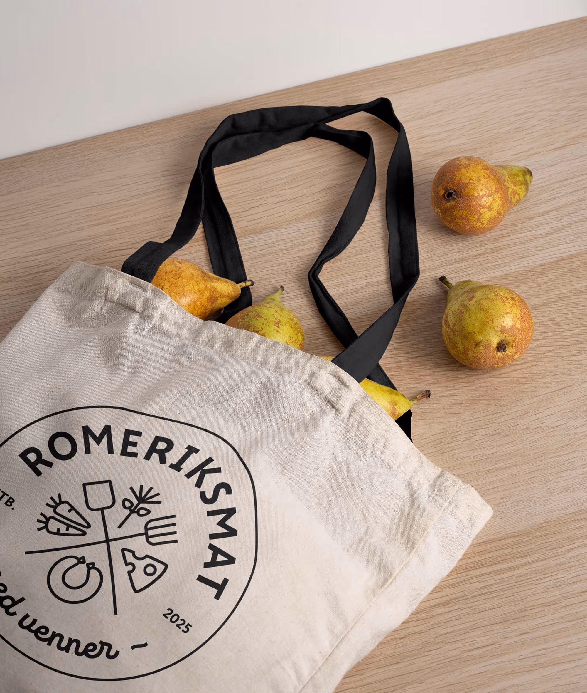 Beige tote bag with black handles and Romeriksmat logo, filled with pears and placed on wooden surface with three pears beside it.
