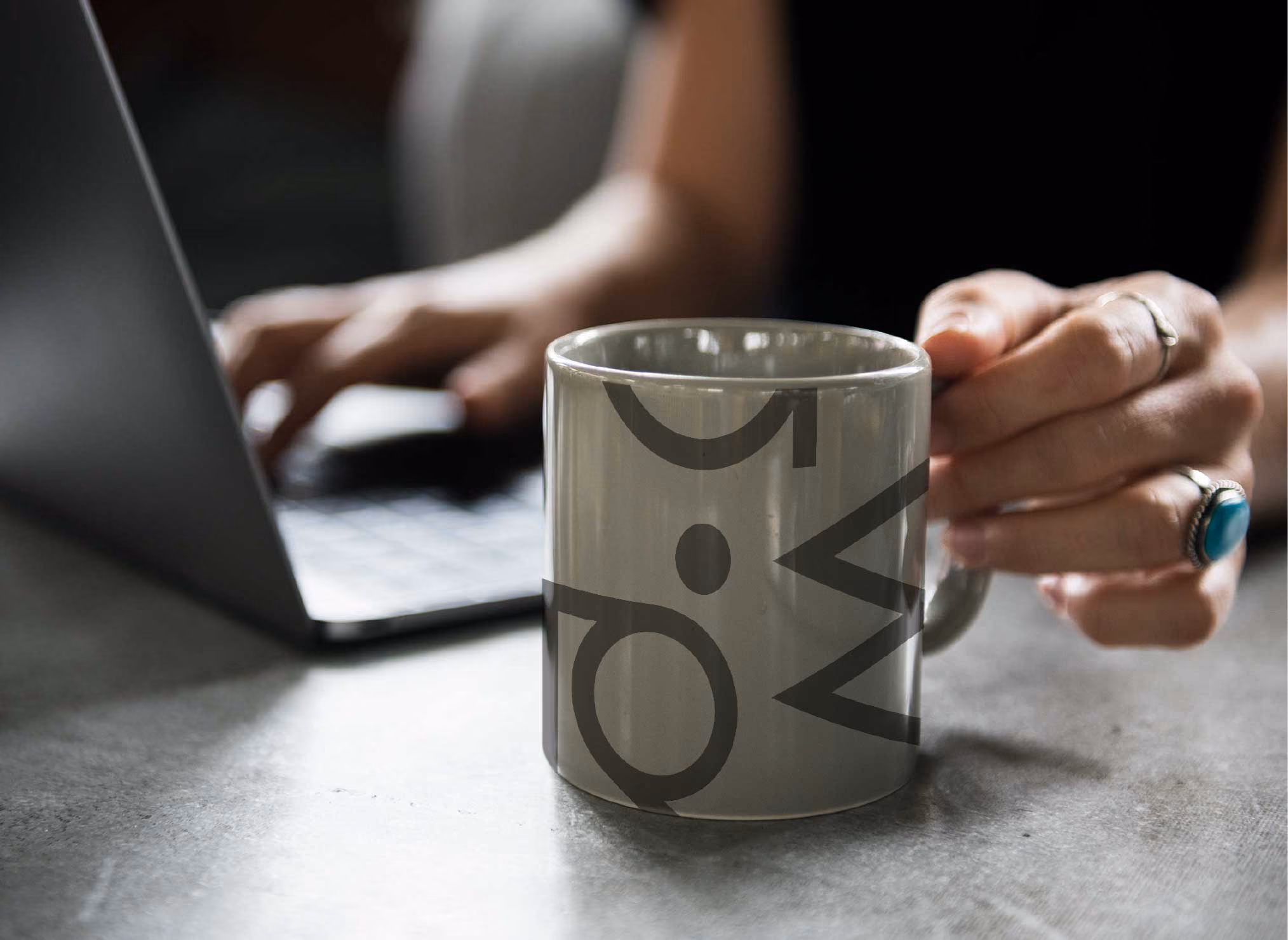 Kopp med logo, hender som jobber på datamaskin i bakgrunnen. RåW studio, Grafisk designer på Jessheim.
