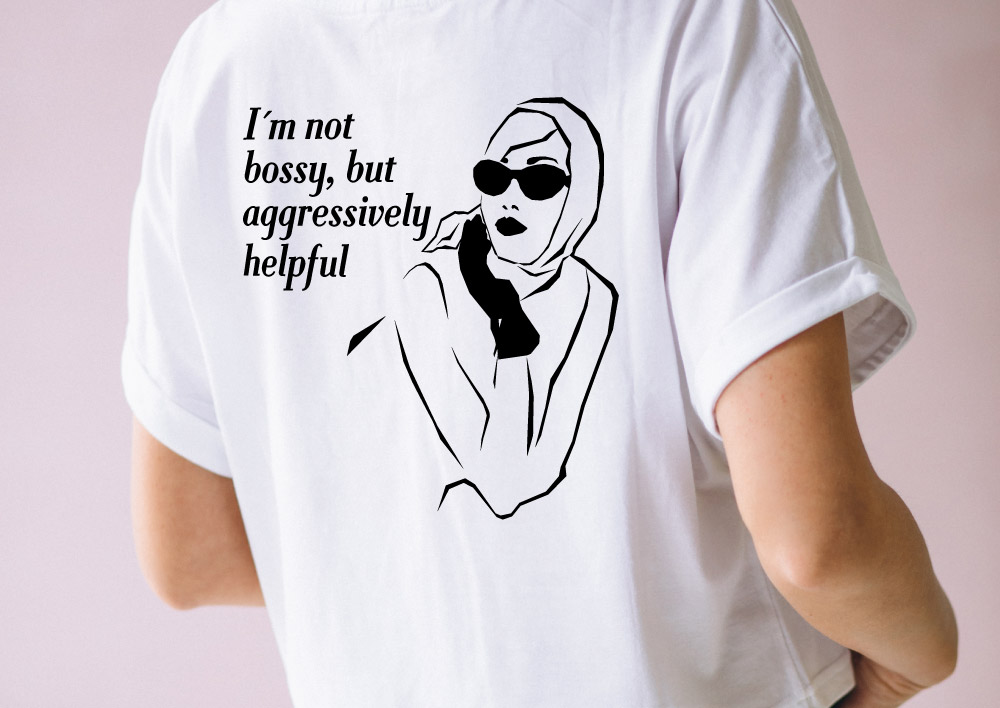 Person wearing a white t-shirt with a black line drawing of a woman wearing sunglasses and a headscarf on the back, next to the text "I'm not bossy, but aggressively helpful."