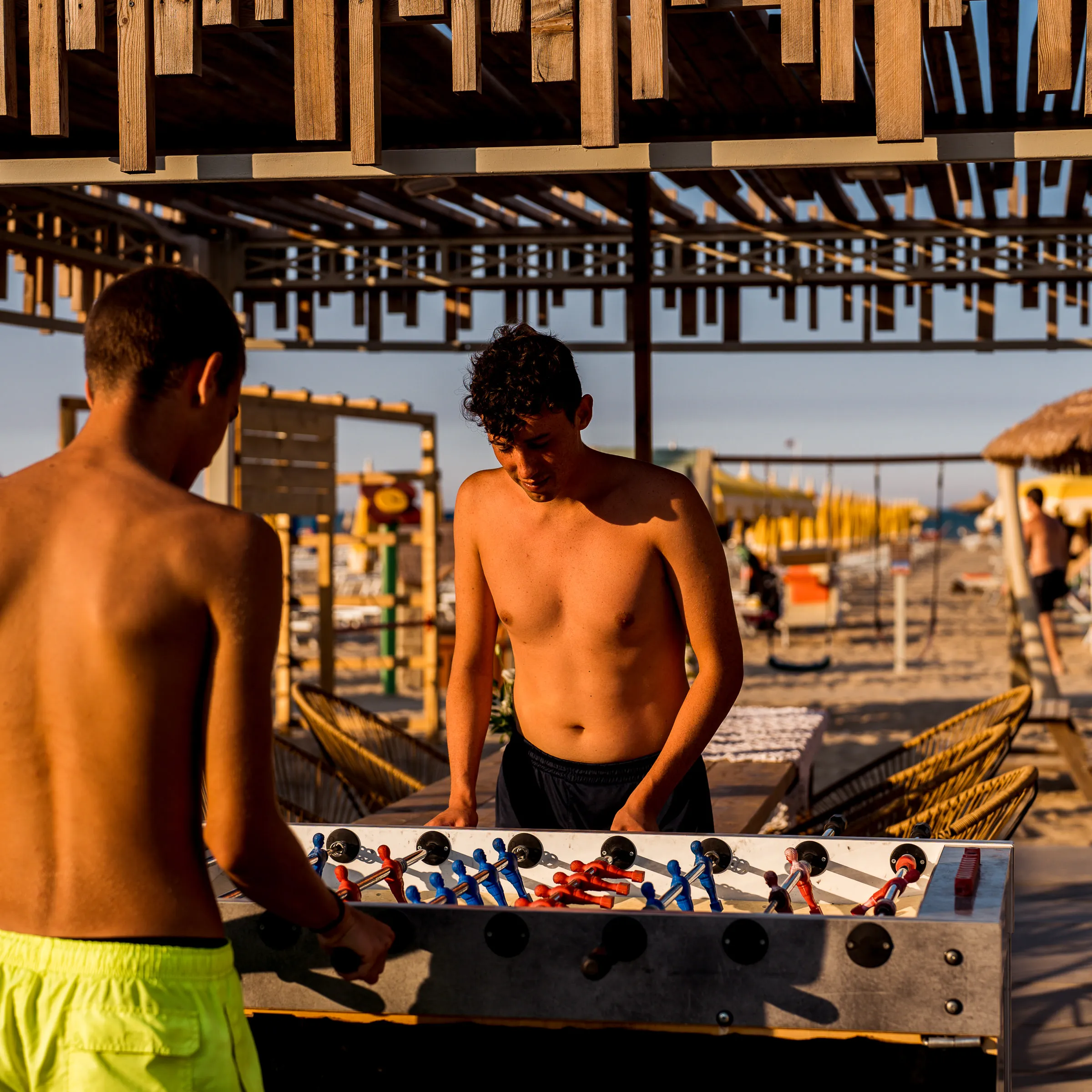 Due ragazzi senza maglietta giocano a calcio balilla all'aperto su una spiaggia al tramonto.