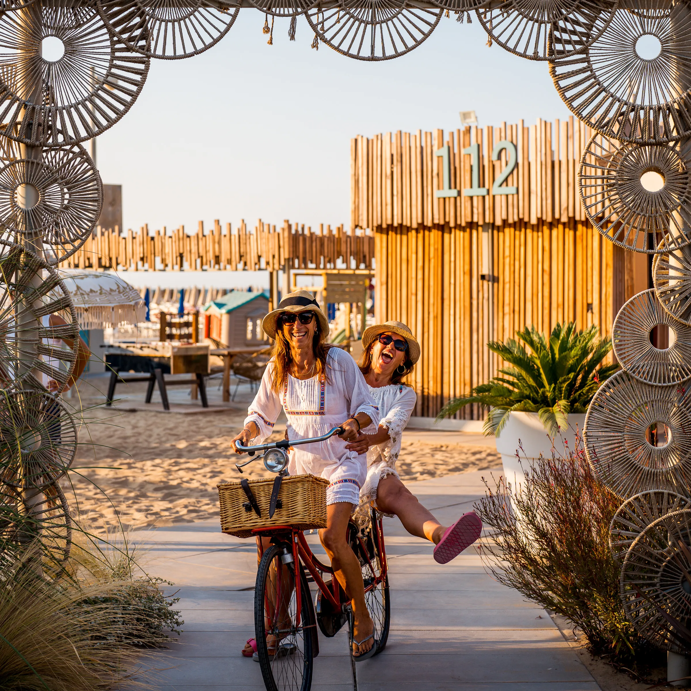Due donne sorridenti in abiti estivi e cappelli che vanno in bicicletta insieme su una passeggiata vicino alla spiaggia al tramonto.