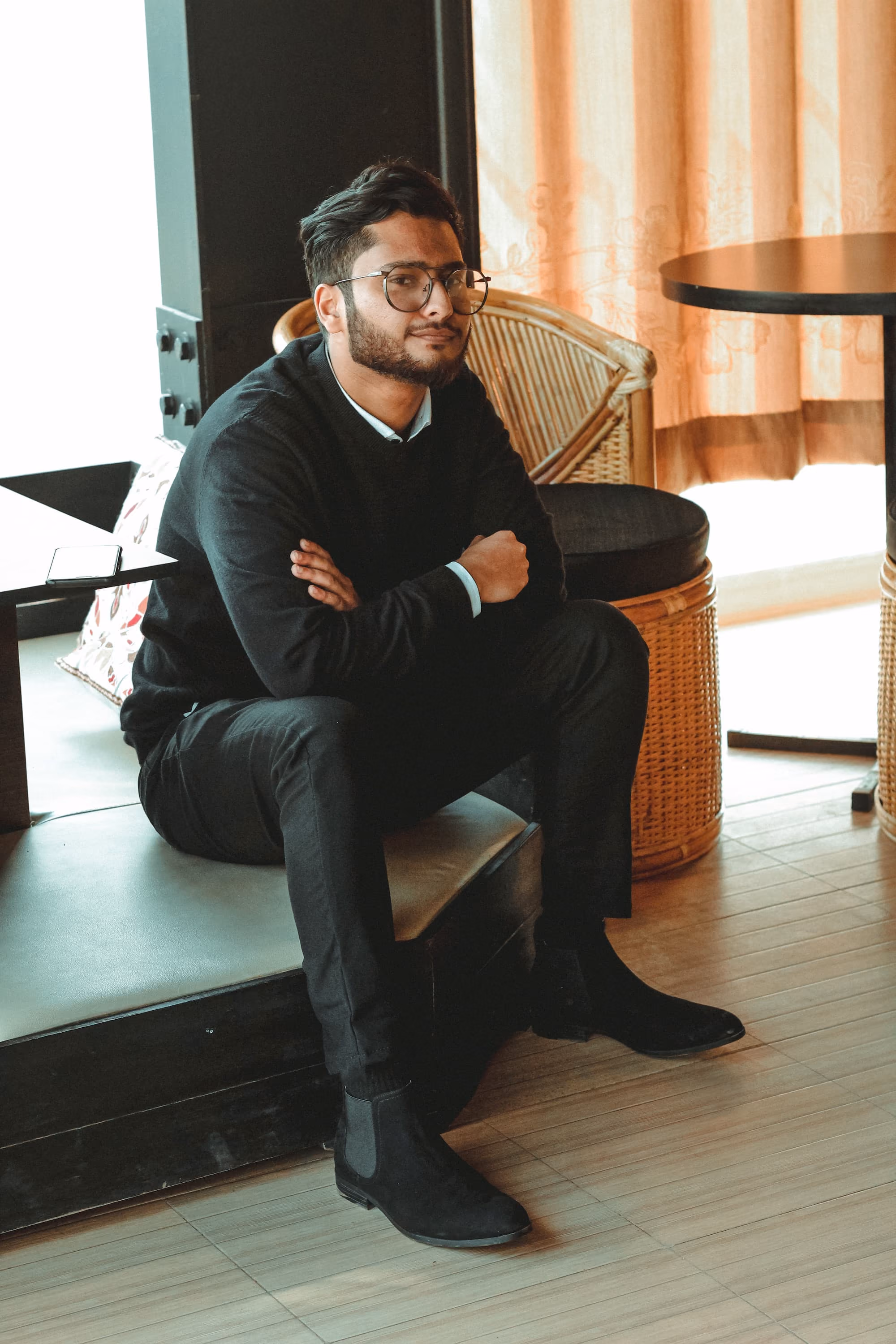 Man wearing glasses and black outfit sitting with arms crossed in a cozy room with wooden floor.