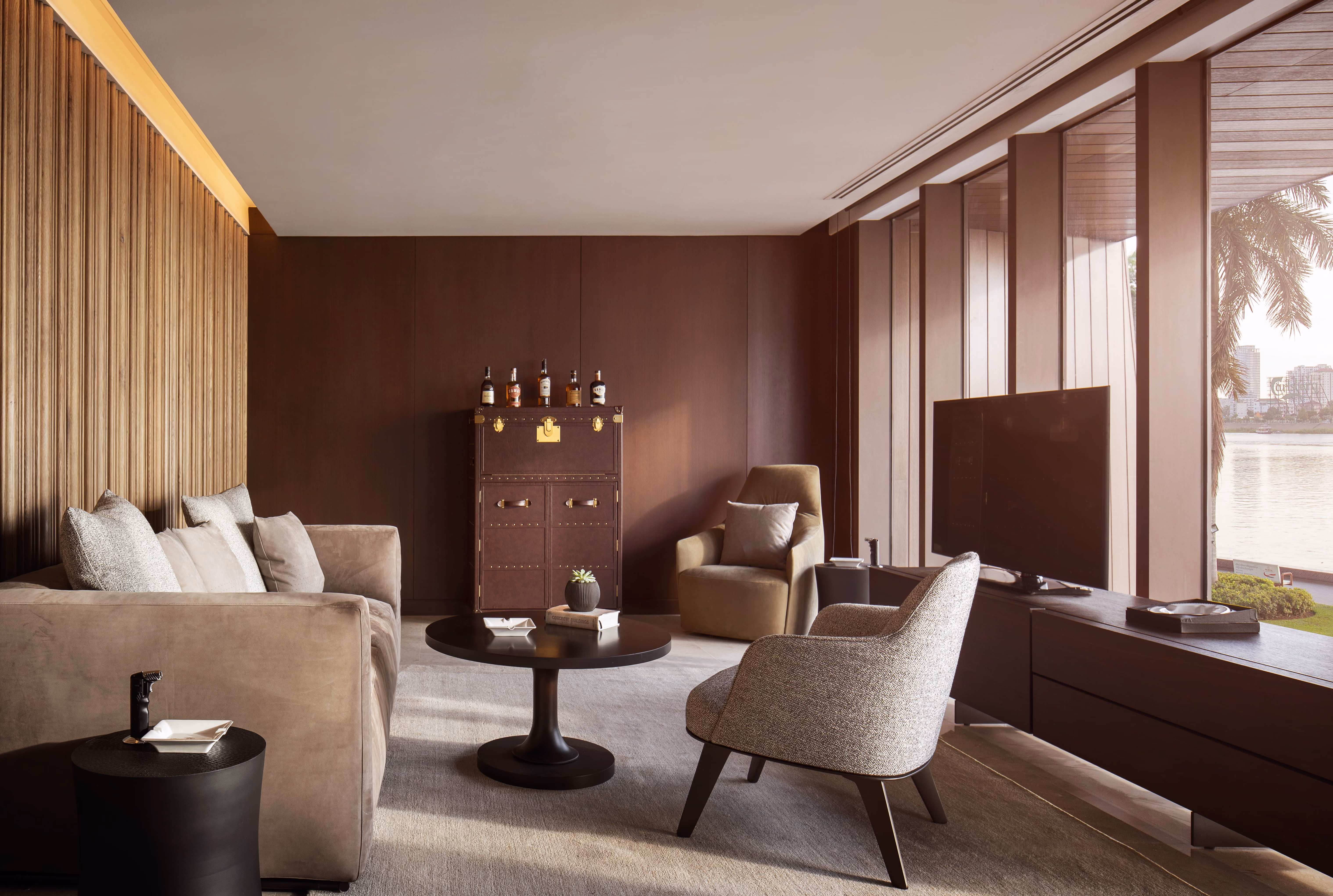 Modern living room with beige sofa, armchairs, round coffee table, and large windows overlooking water.