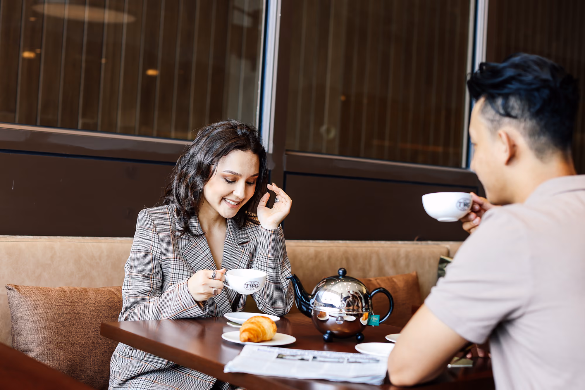 A woman and a man enjoying tea with a croissant and newspaper at a cafe table.