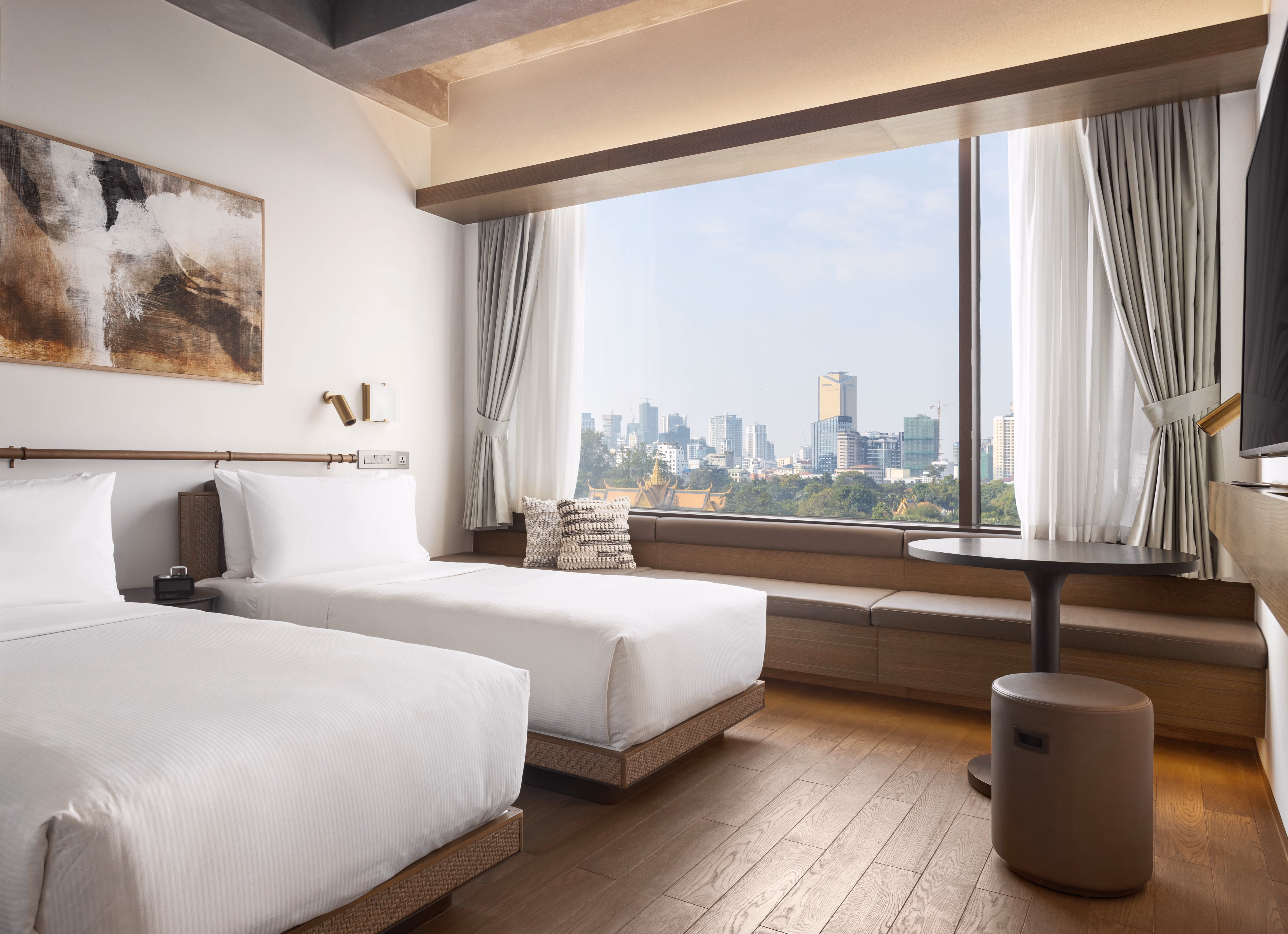 Hotel room with two white twin beds, city view window, bench seating, and round table with stool.