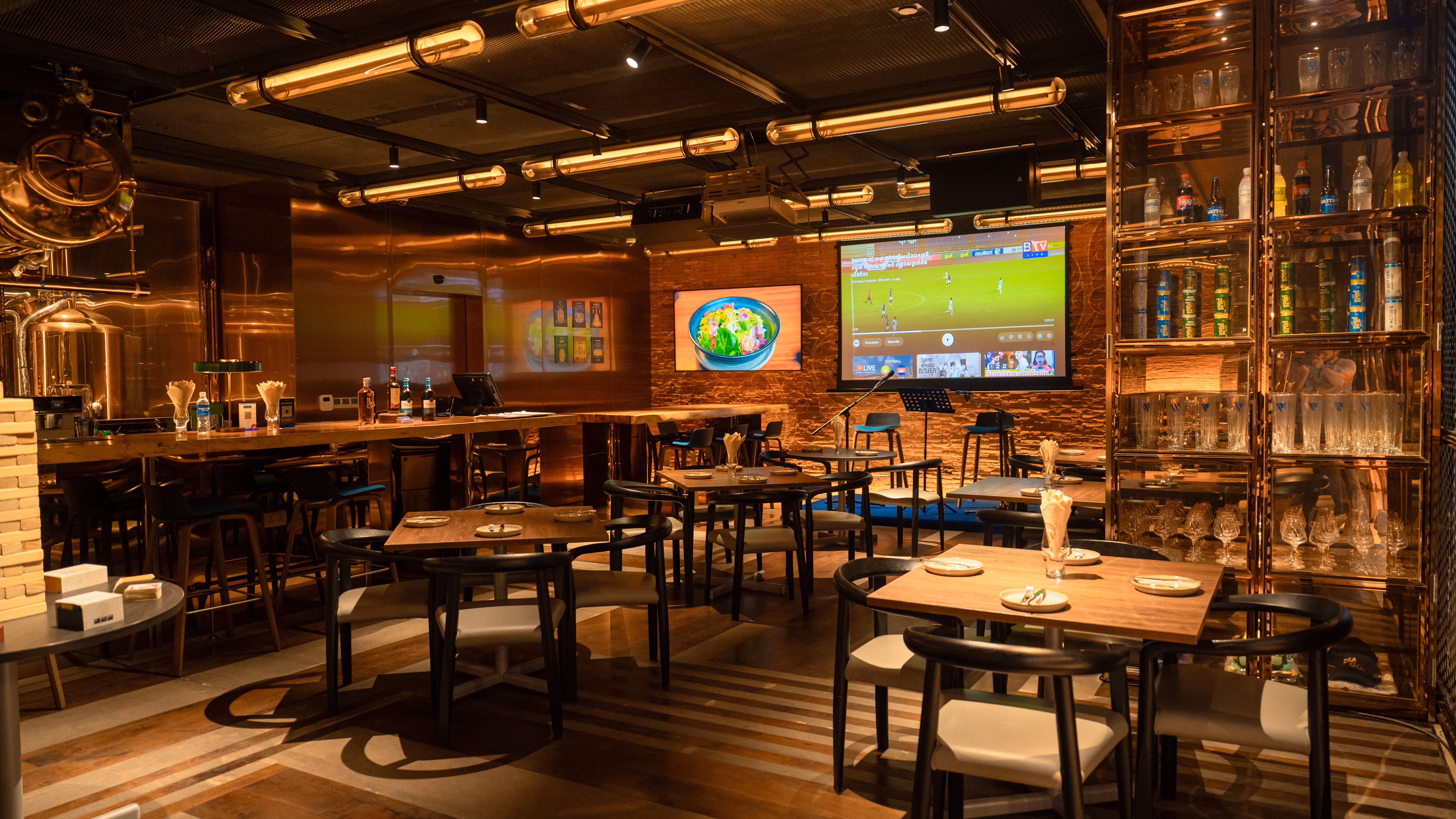 Warm-lit restaurant interior with wooden tables, chairs, bar, and two screens on brick wall.