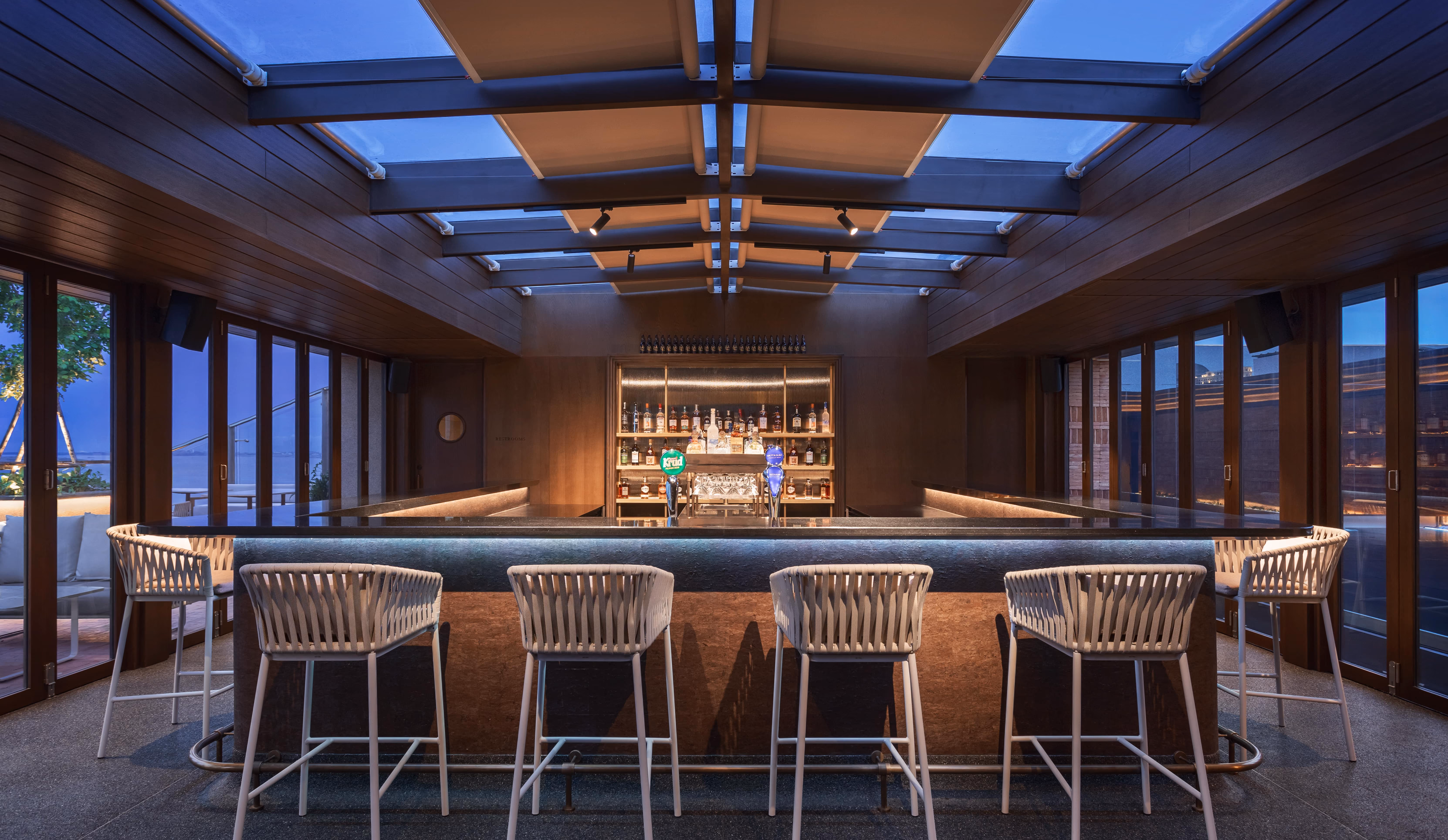 Modern bar with six white rope chairs, glass roof panels, and a stocked liquor shelf behind the counter.