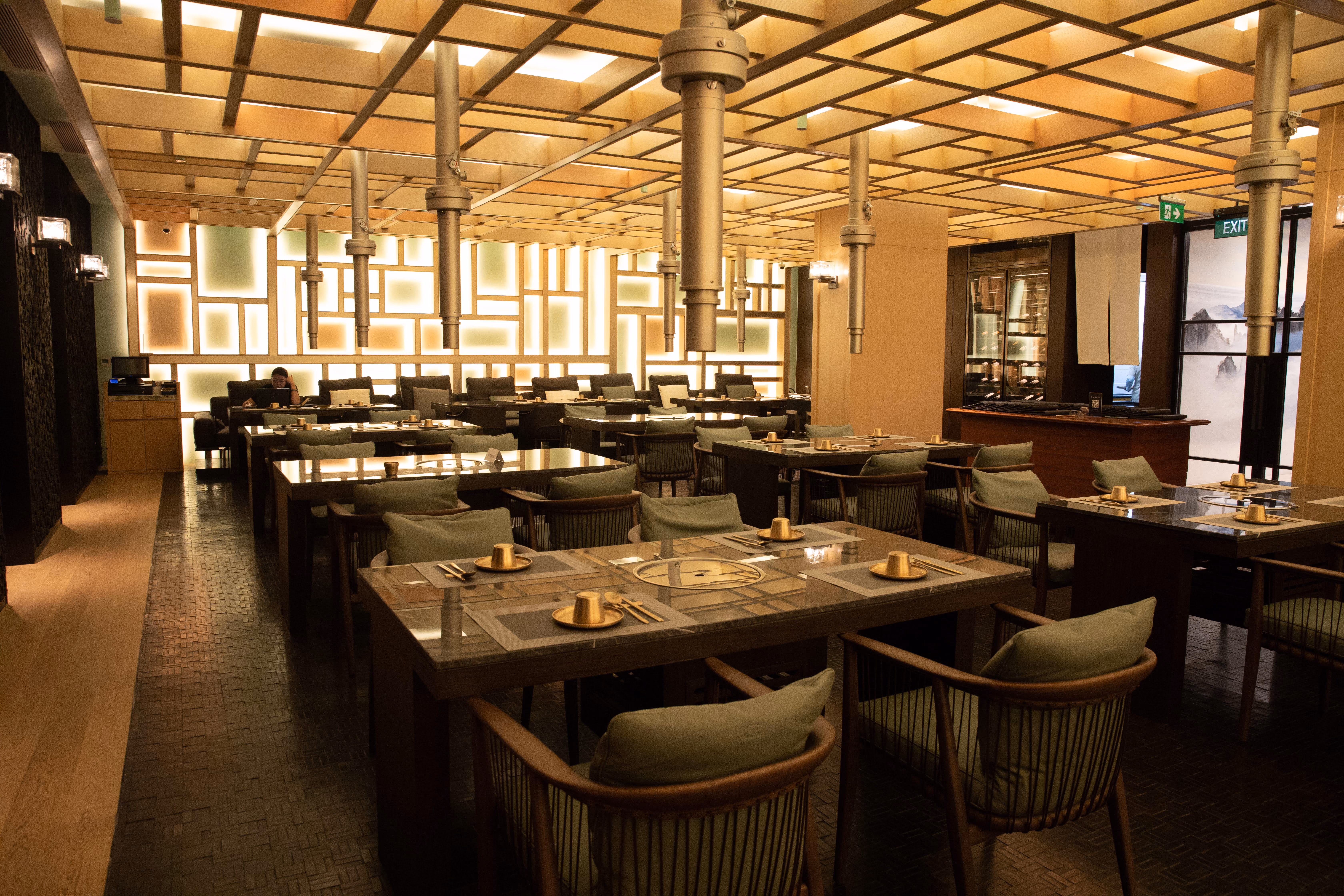 Modern restaurant interior with wooden tables, green cushioned chairs, and geometric backlit wall panels.