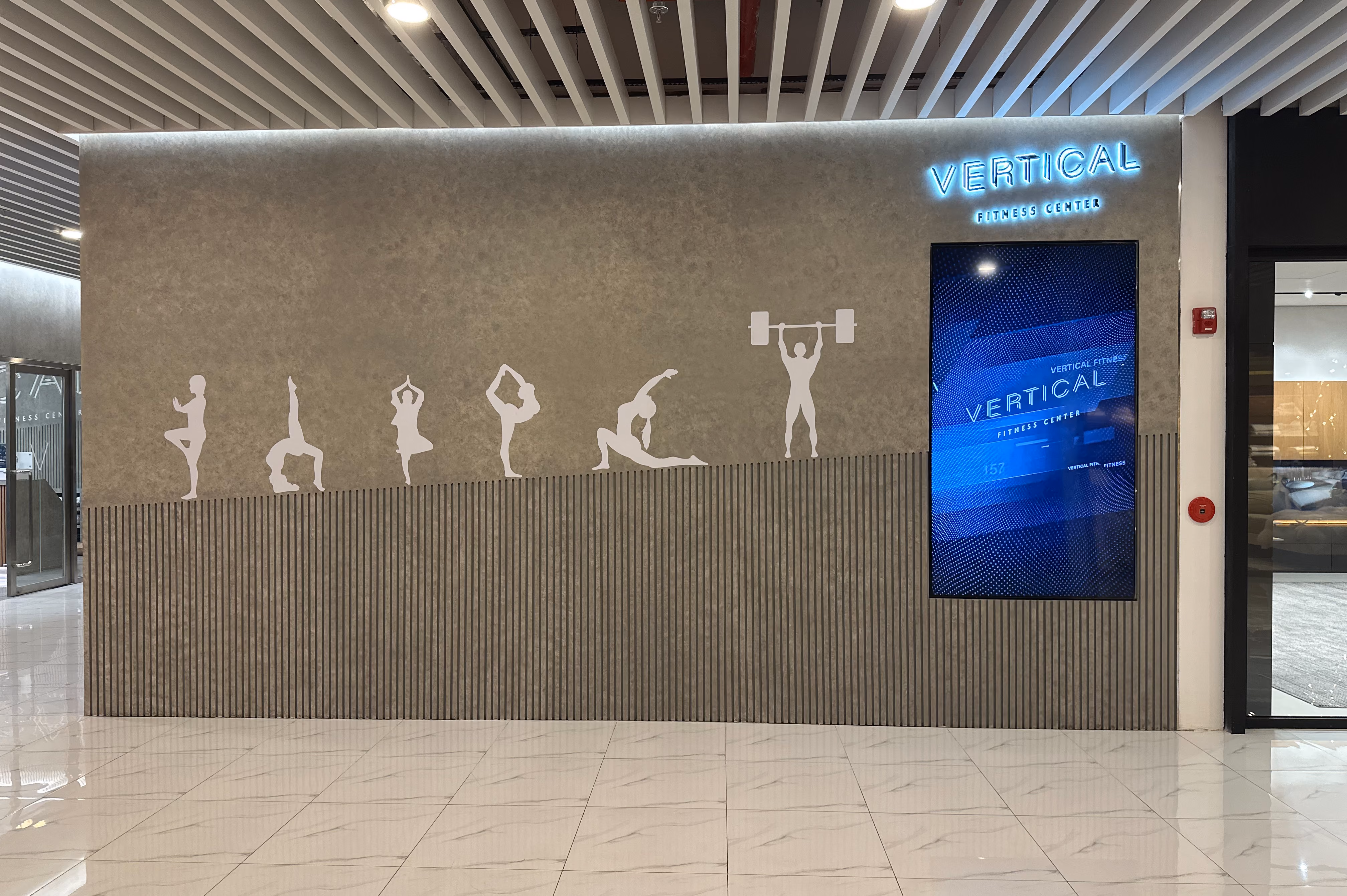 Gym entrance wall with fitness silhouettes and a digital screen displaying Vertical Fitness Center signage.