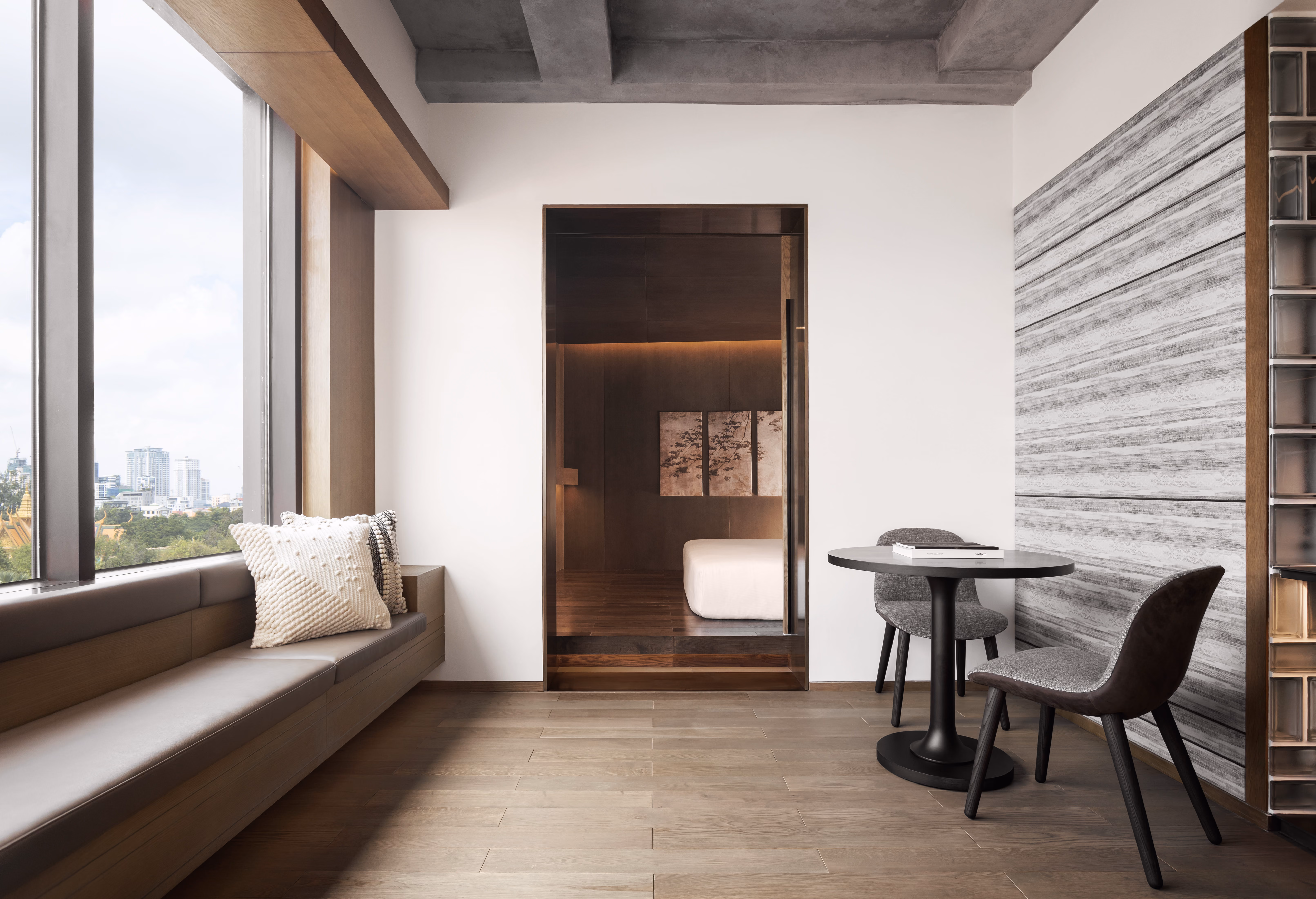 Modern hotel room seating area with window bench, two chairs, round table, and view into bedroom.