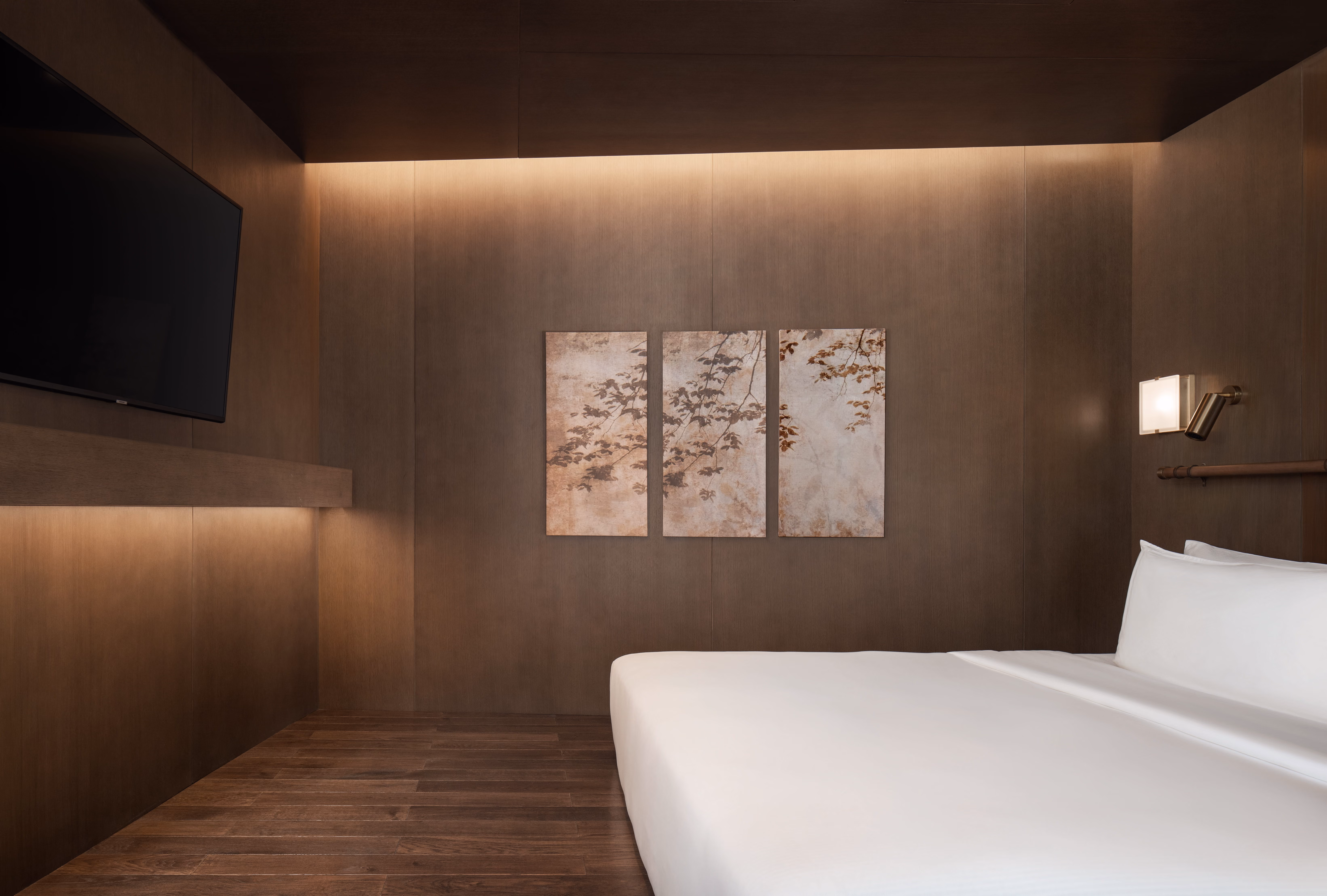 Minimalist bedroom with large bed, wall art of tree branches, wooden floor, and mounted TV on wood panel wall.