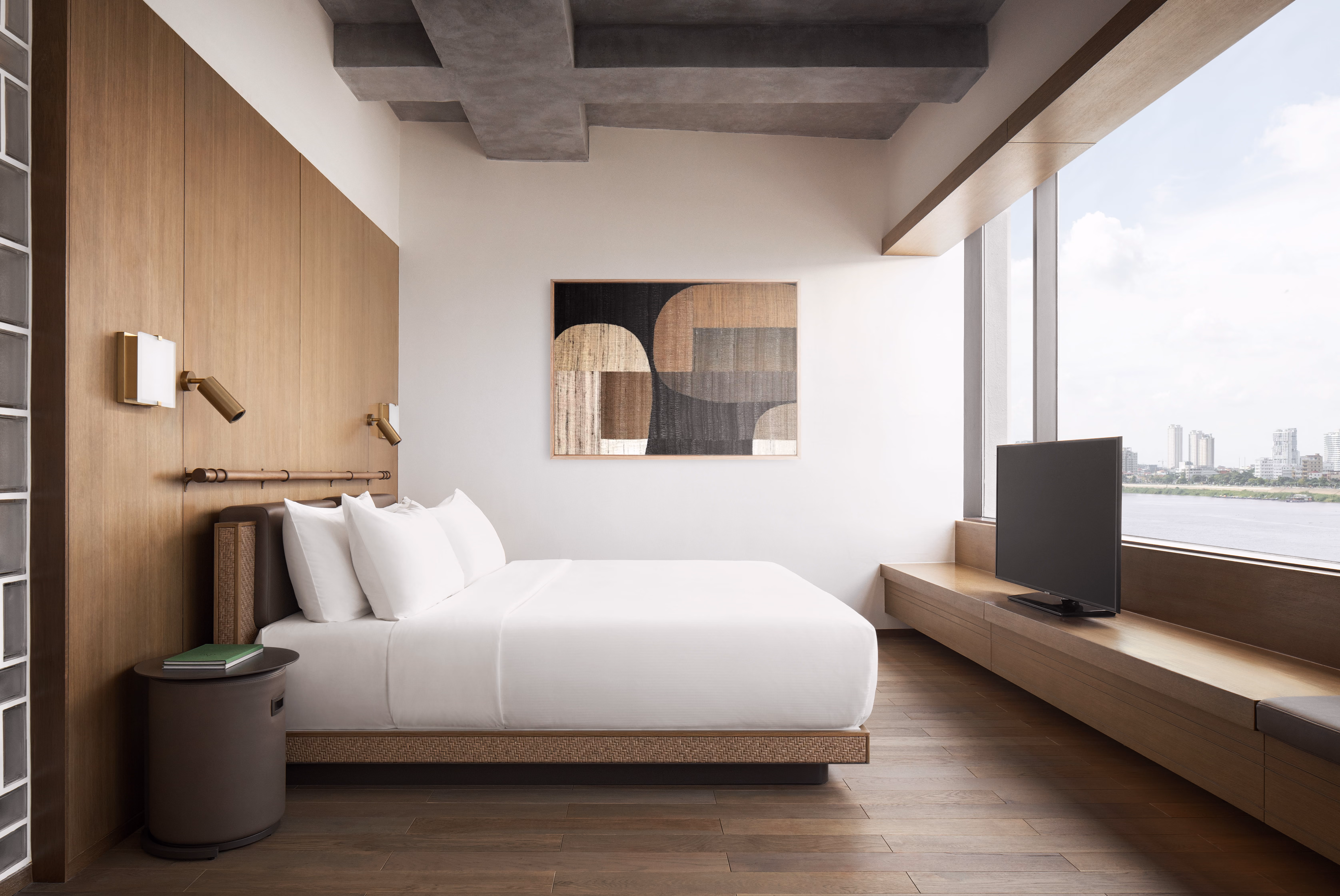 Minimalist hotel room with white bed, wooden panel wall, abstract art, and river view from large window.