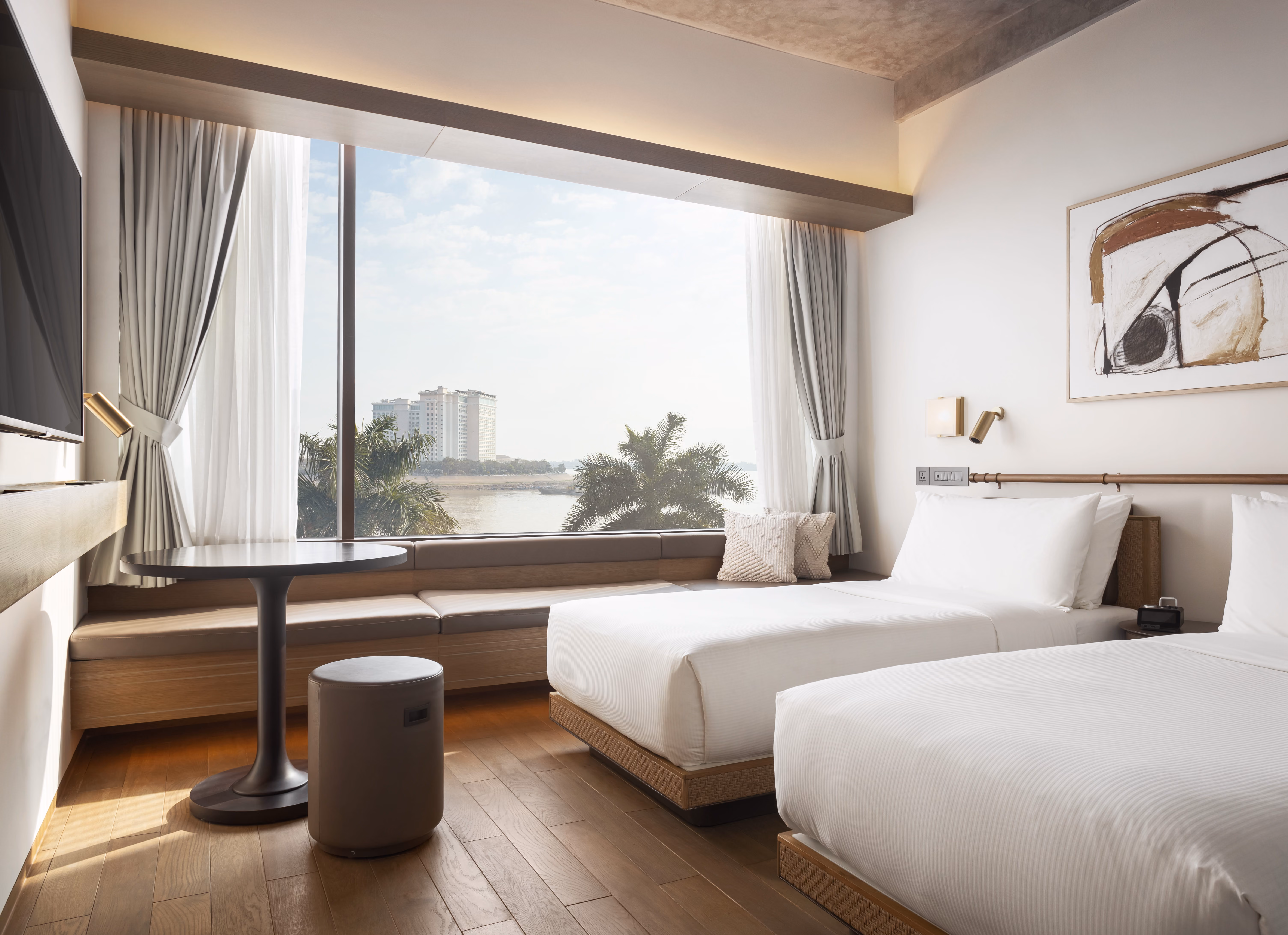 Modern hotel room with two beds, large window with river view, bench seating, and round table with stool.