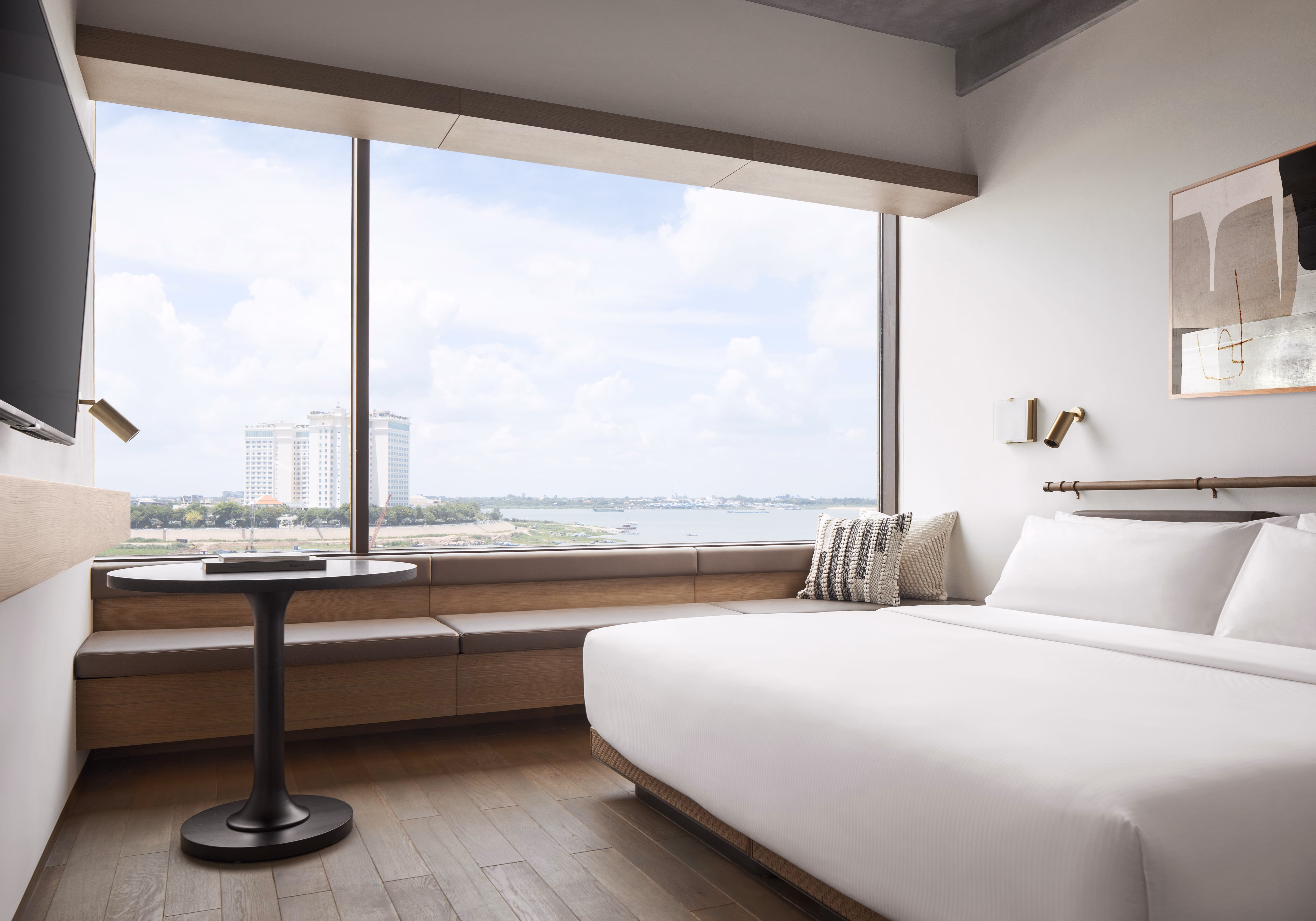 Modern hotel room with large window seat, round table, king bed, and view of city and river.