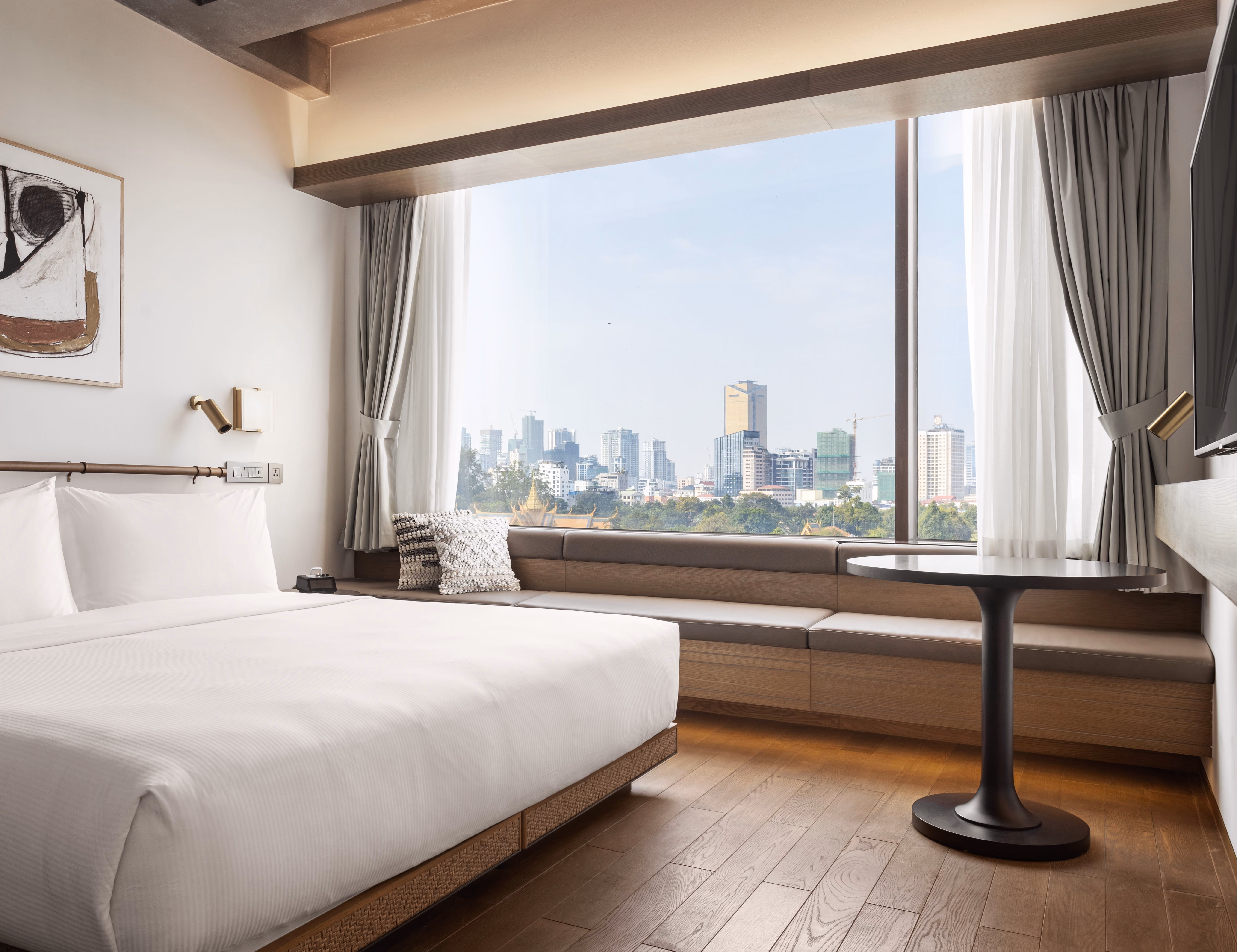 Modern hotel room with king bed, large window seat, round table, and city skyline view outside.