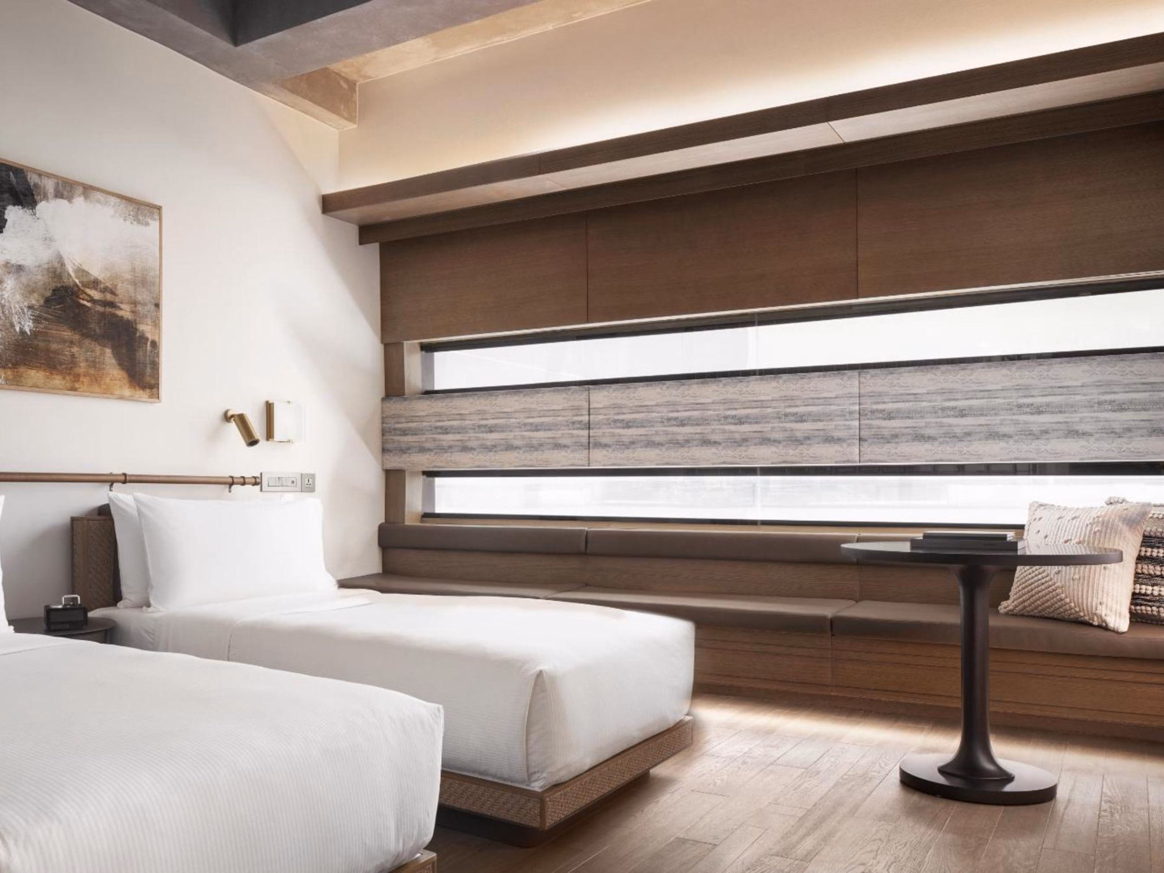 Modern hotel room with two white beds, a long wood bench, round table, and abstract wall art.
