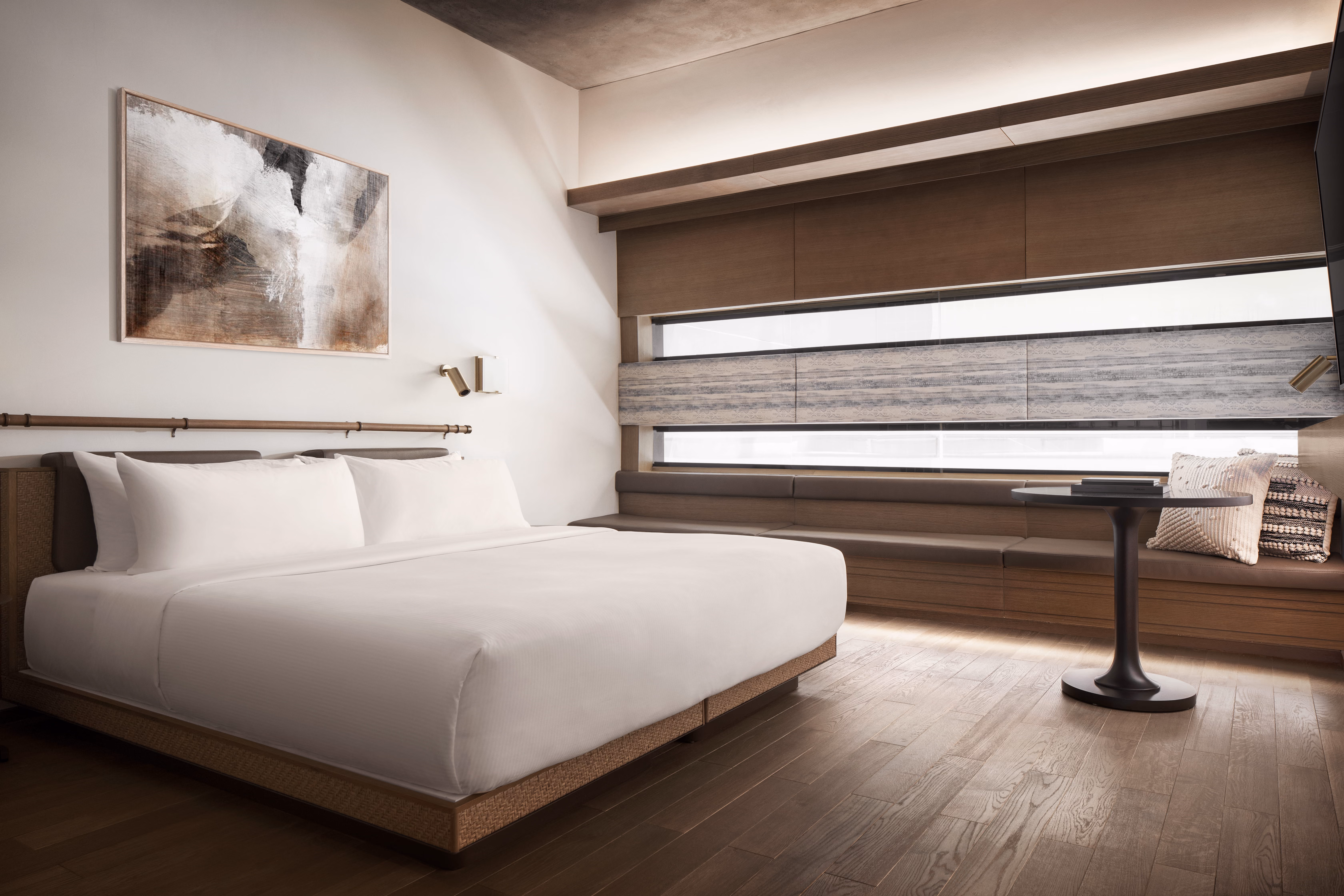 Modern hotel room with a king bed, abstract wall art, long bench, pillows, and round table.
