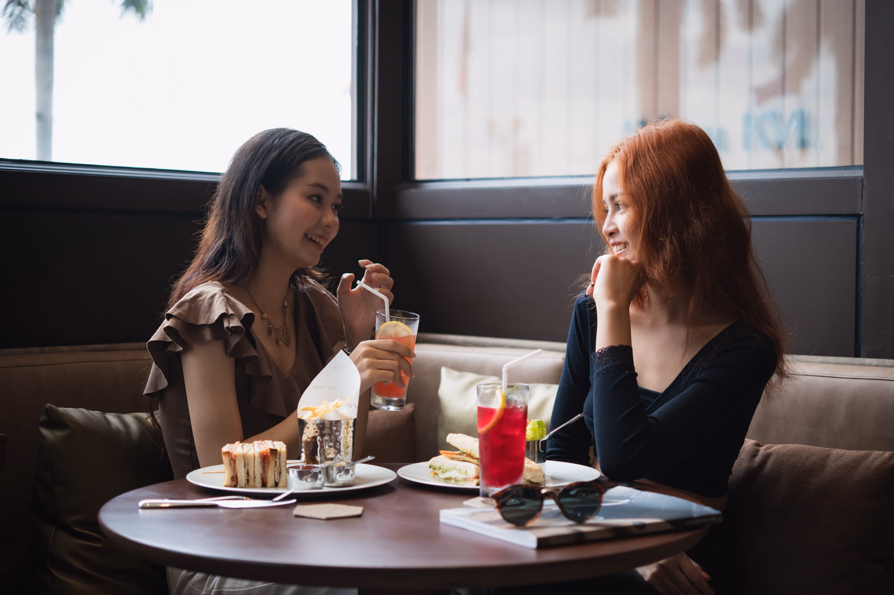 Two women smiling and talking while sitting at a table with sandwiches, fries, and drinks.