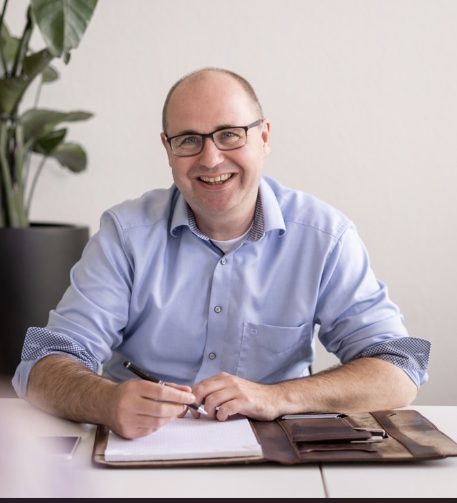 Blije man met bril en blauw overhemd schrijft in een notitieboekje aan een bureau met een grote plantenpot op de achtergrond.