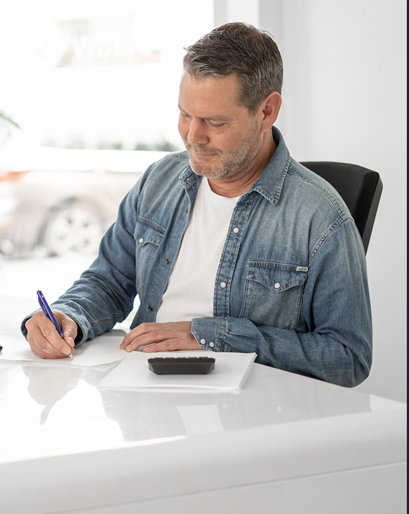 Man schrijft met pen op papier aan een witte tafel, met een rekenmachine voor hem.