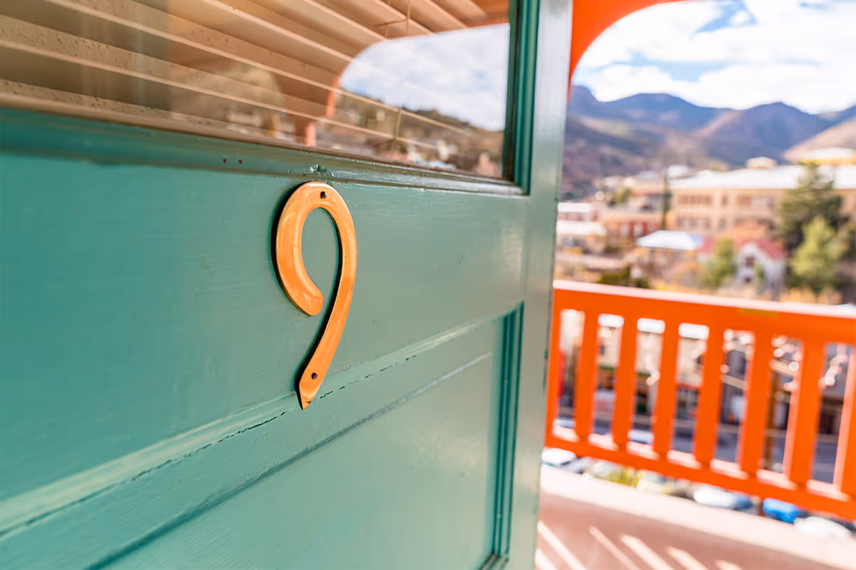 Close-up of an orange number 9 on a teal door with a blurred outdoor balcony and mountains in the background.