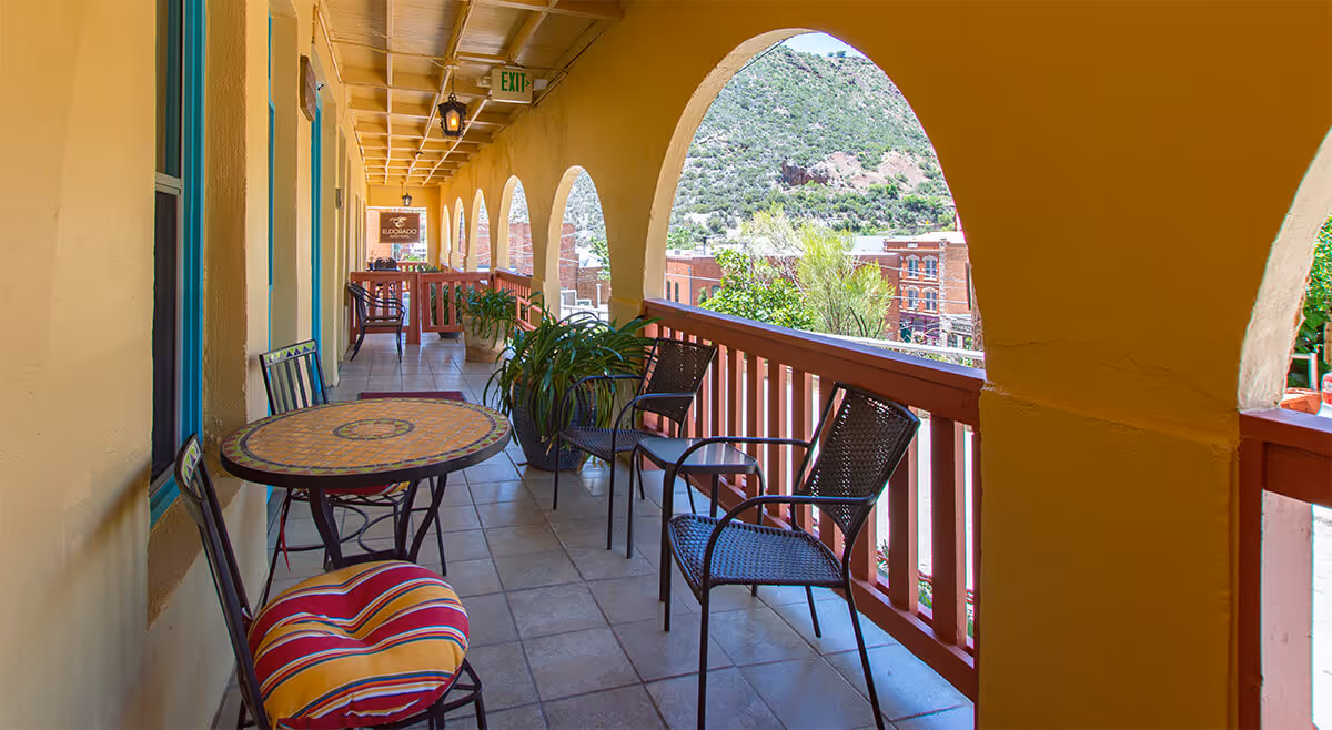 Covered balcony with tiled floor, yellow walls, arches, metal chairs, tables, and potted plants overlooking a green hillside and brick buildings.