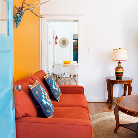 Cozy living room with a red sofa, patterned cushions, wooden coffee table, and a side table with a colorful lamp under bright natural light.