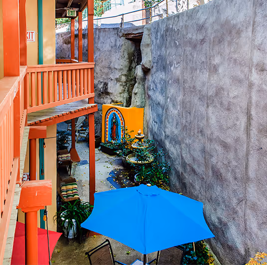 Outdoor patio area with orange wooden railings, a blue umbrella, chairs, a water fountain, and a colorful religious mural on a yellow wall.
