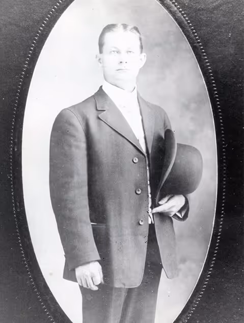 Black and white portrait of a man in a suit holding a hat, framed in an oval border.