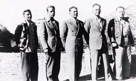 Black and white photo of five men standing outdoors in a line, dressed in mid-20th century suits and jackets.