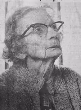 Black and white photo of an elderly woman with cat-eye glasses, wearing a textured sweater and looking upward.
