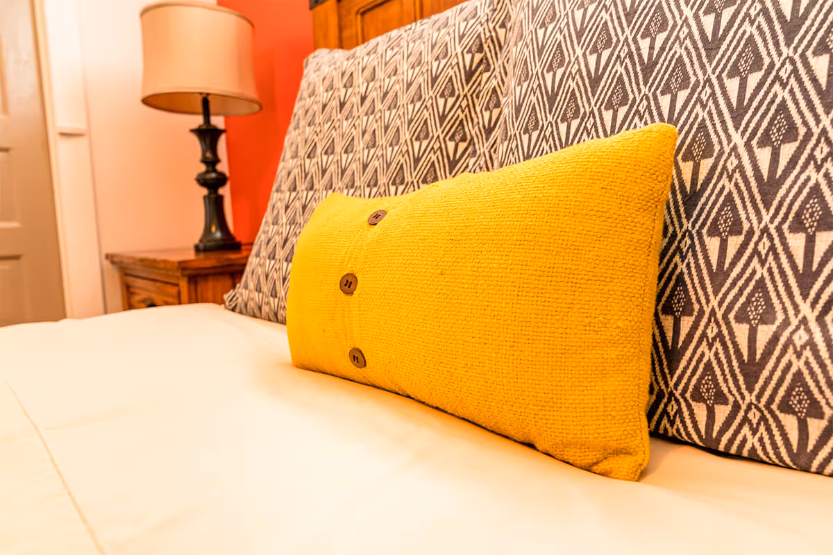 Close-up of a bed with a yellow buttoned pillow and a patterned headboard, next to a wooden nightstand with a lamp.
