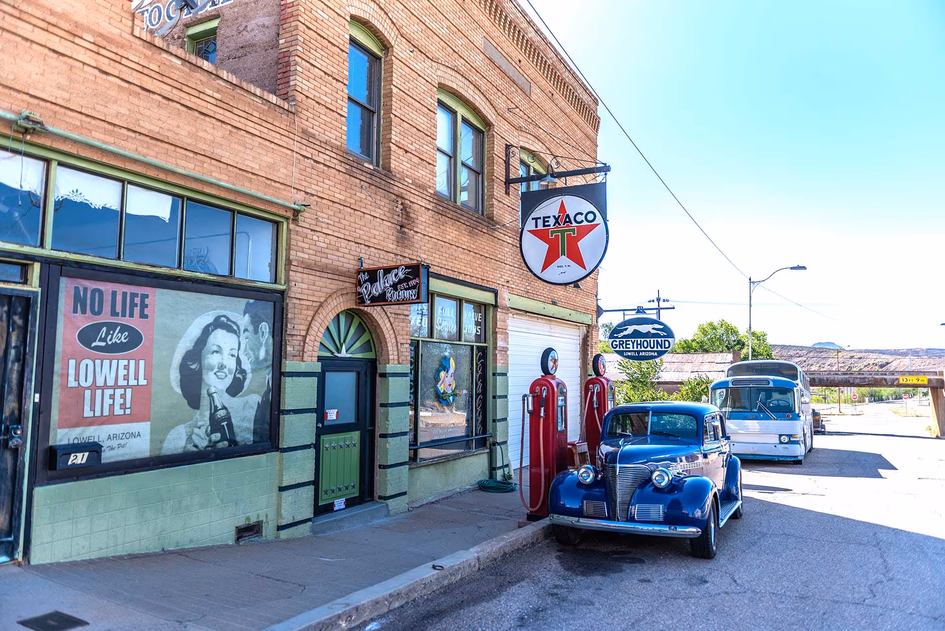 Vintage blue car parked beside red Texaco gas pumps at a brick building with Lowe Arizona Greyhound sign and retro Lowell life poster.