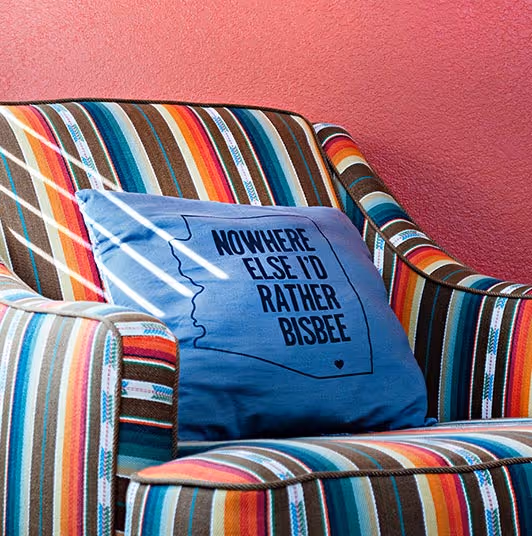 Striped armchair with a blue pillow that reads 'Nowhere else I'd rather Bisbee' against a pink wall.