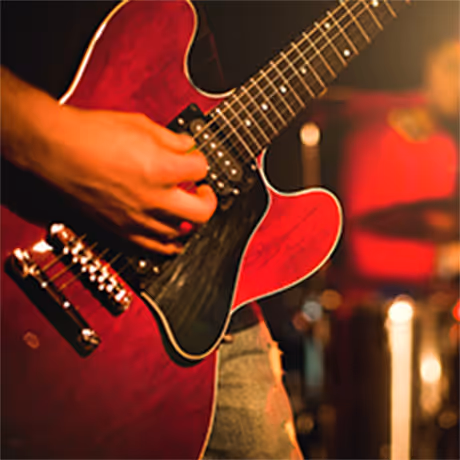 Close-up of a person playing a red electric guitar on stage with blurred drummer in the background.