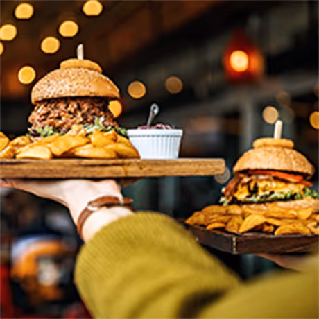 Two wooden trays each carrying a burger with fries and a small bowl of sauce, held by hands in a restaurant setting with blurred lights in the background.