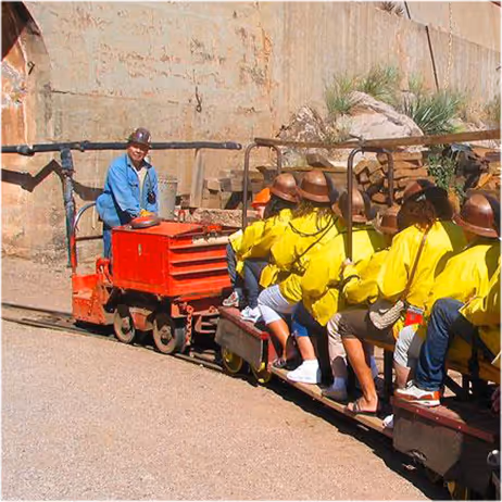 Group of people wearing yellow jackets and hats riding a small open train led by a person in a blue jacket and helmet.