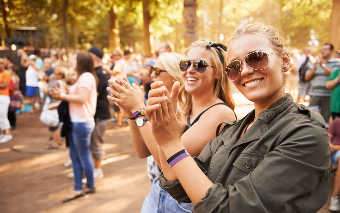 Two young women wearing sunglasses smiling and clapping at an outdoor event with a crowd and trees in the background.