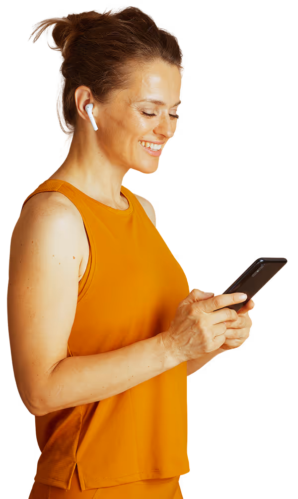 Smiling woman wearing wireless earbuds and an orange sleeveless top, holding and looking at a smartphone.