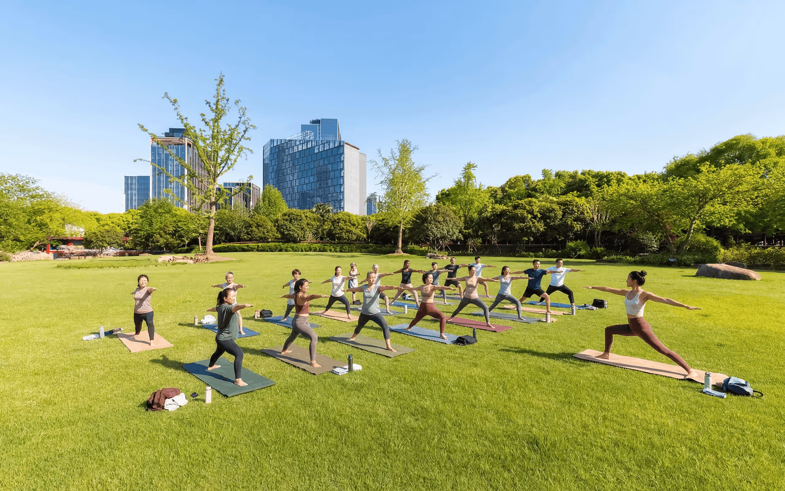 Group of people practicing yoga on mats in a sunny city park with buildings and trees in the background.