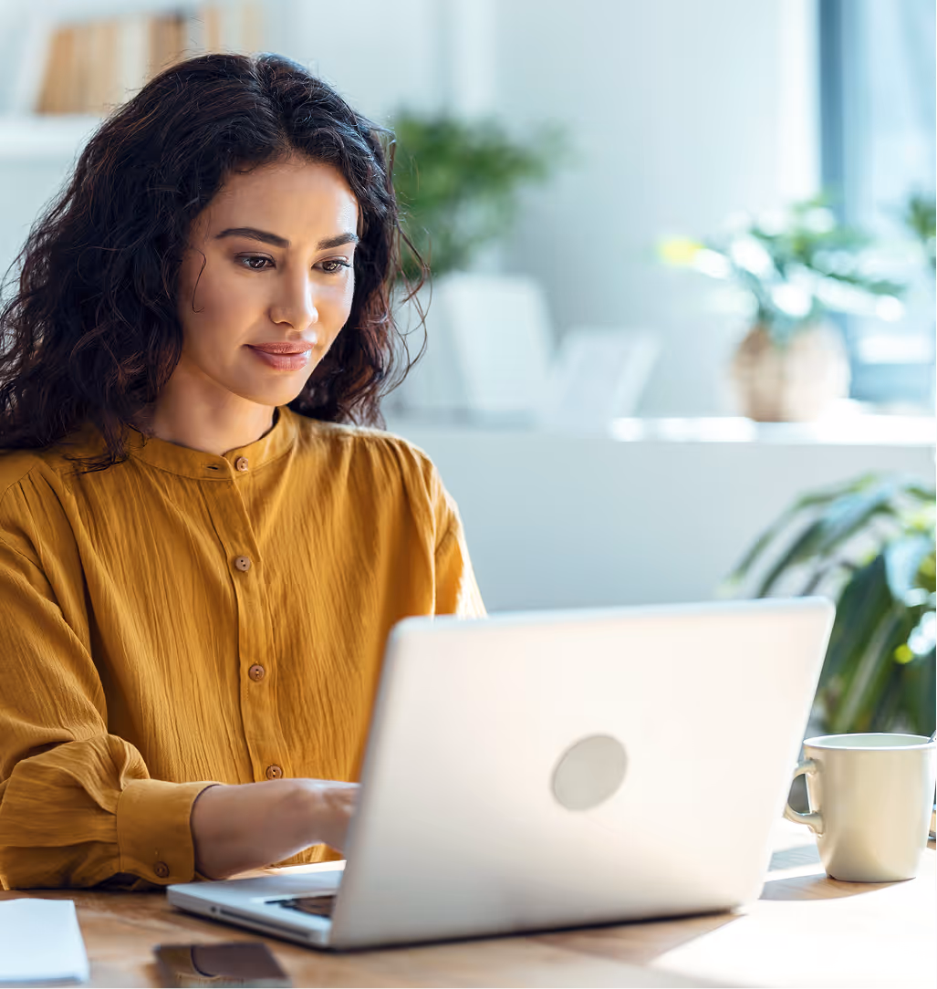 Woman in mustard blouse working on a laptop at a wooden desk with a mug and plants nearby.