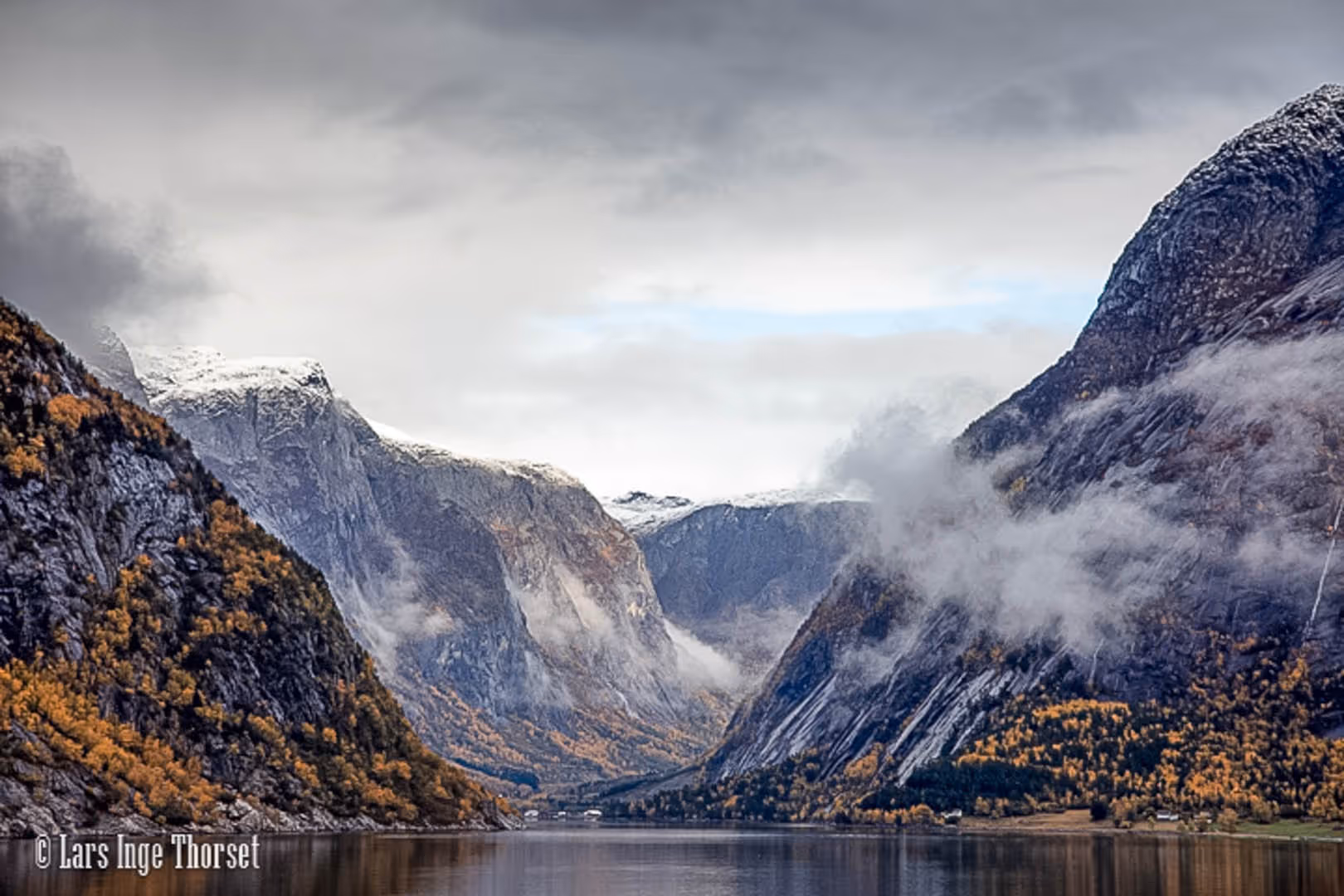 Calm fjord surrounded by steep rocky mountains with autumn foliage and snow-capped peaks under cloudy sky.