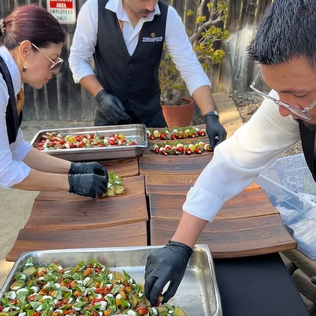 Three staff members are preparing appetizers on wooden plates outside.