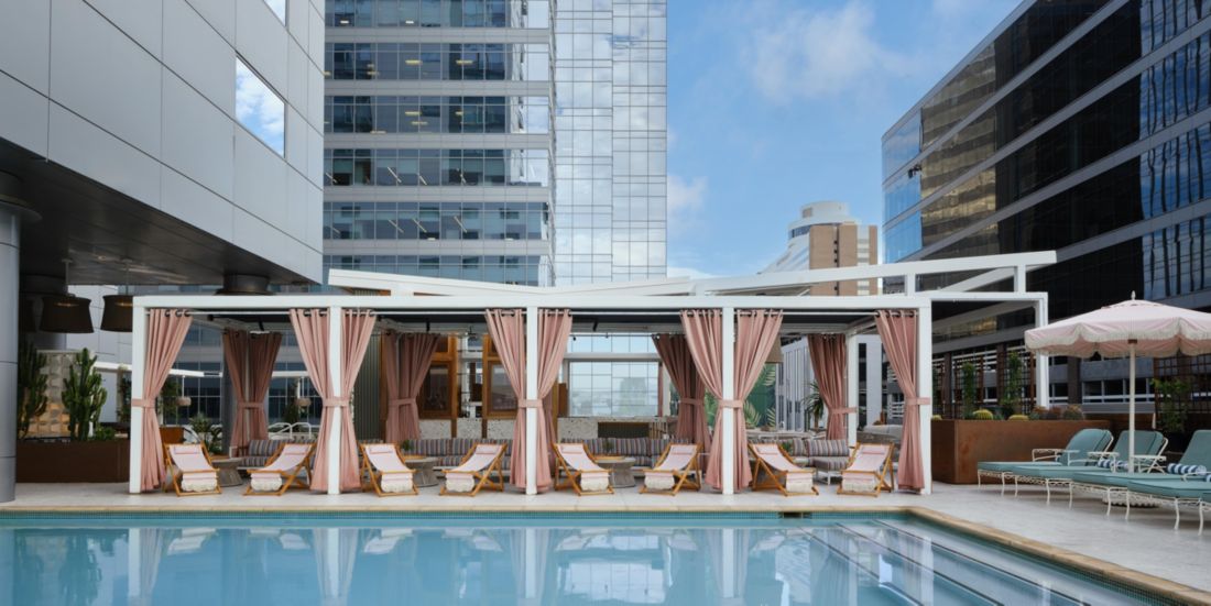 Outdoor pool area with lounge chairs and cabanas draped in pink curtains among tall city skyscrapers.